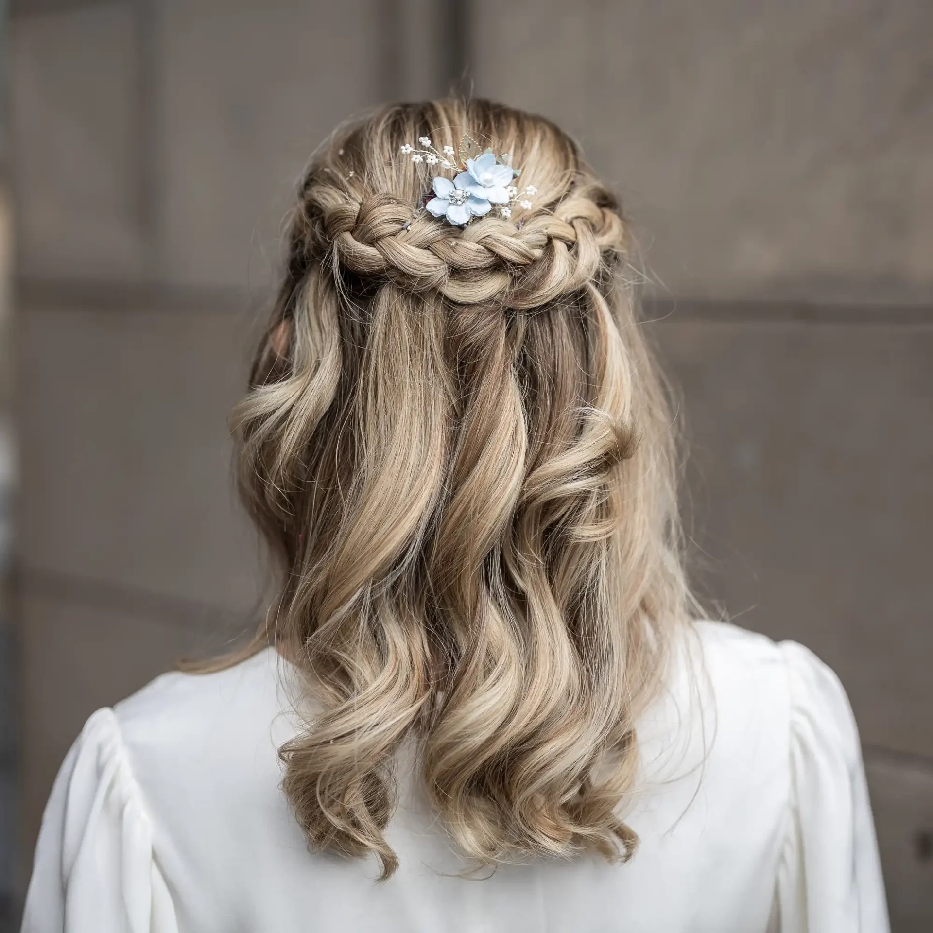 Blonde hair styled in braided crown with loose curls and small blue flowers, seen from the back.