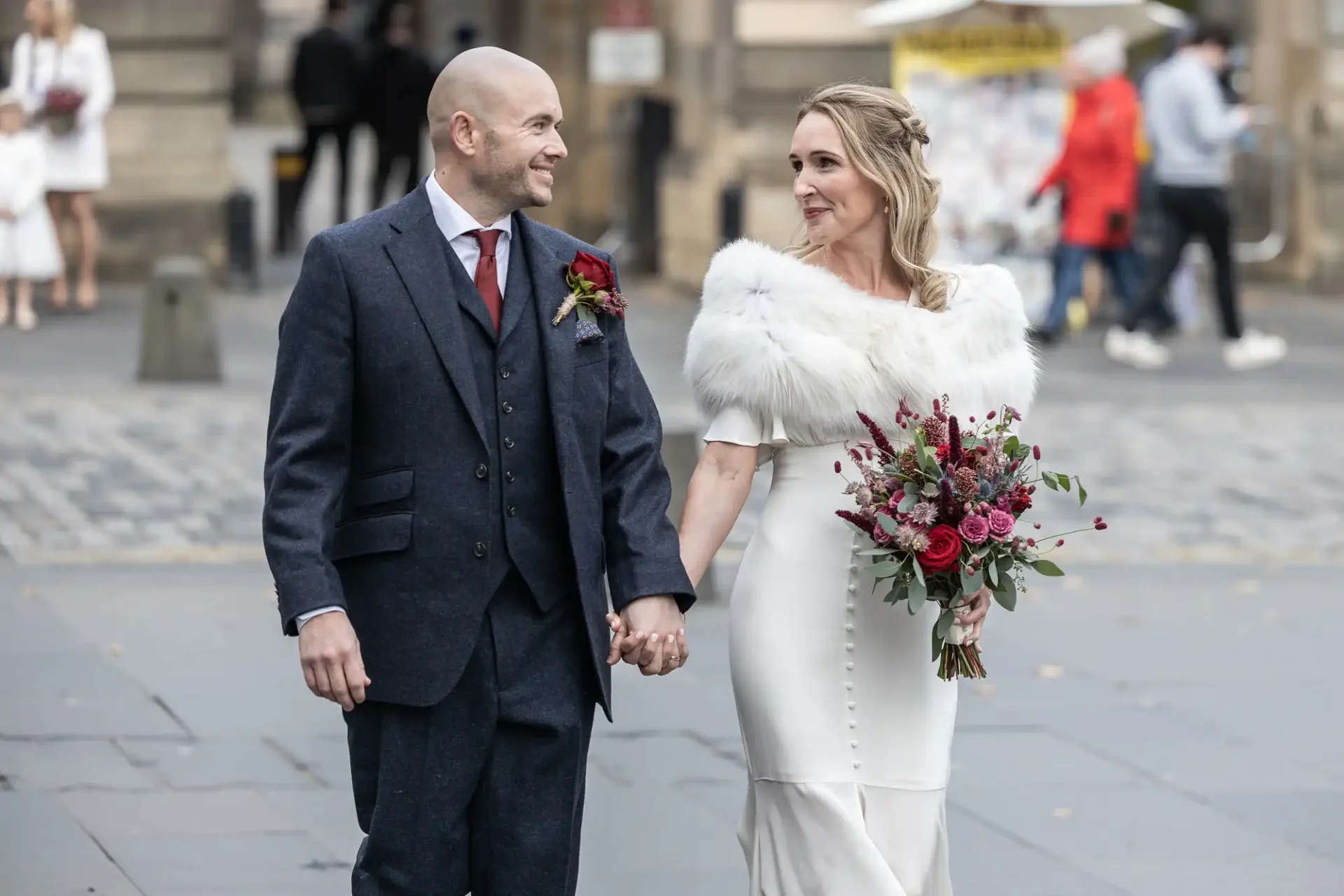 A couple in formal attire walks hand in hand on a city street. The woman holds a bouquet of red flowers, wearing a white dress with a fur stole. The man wears a dark suit with a red tie.