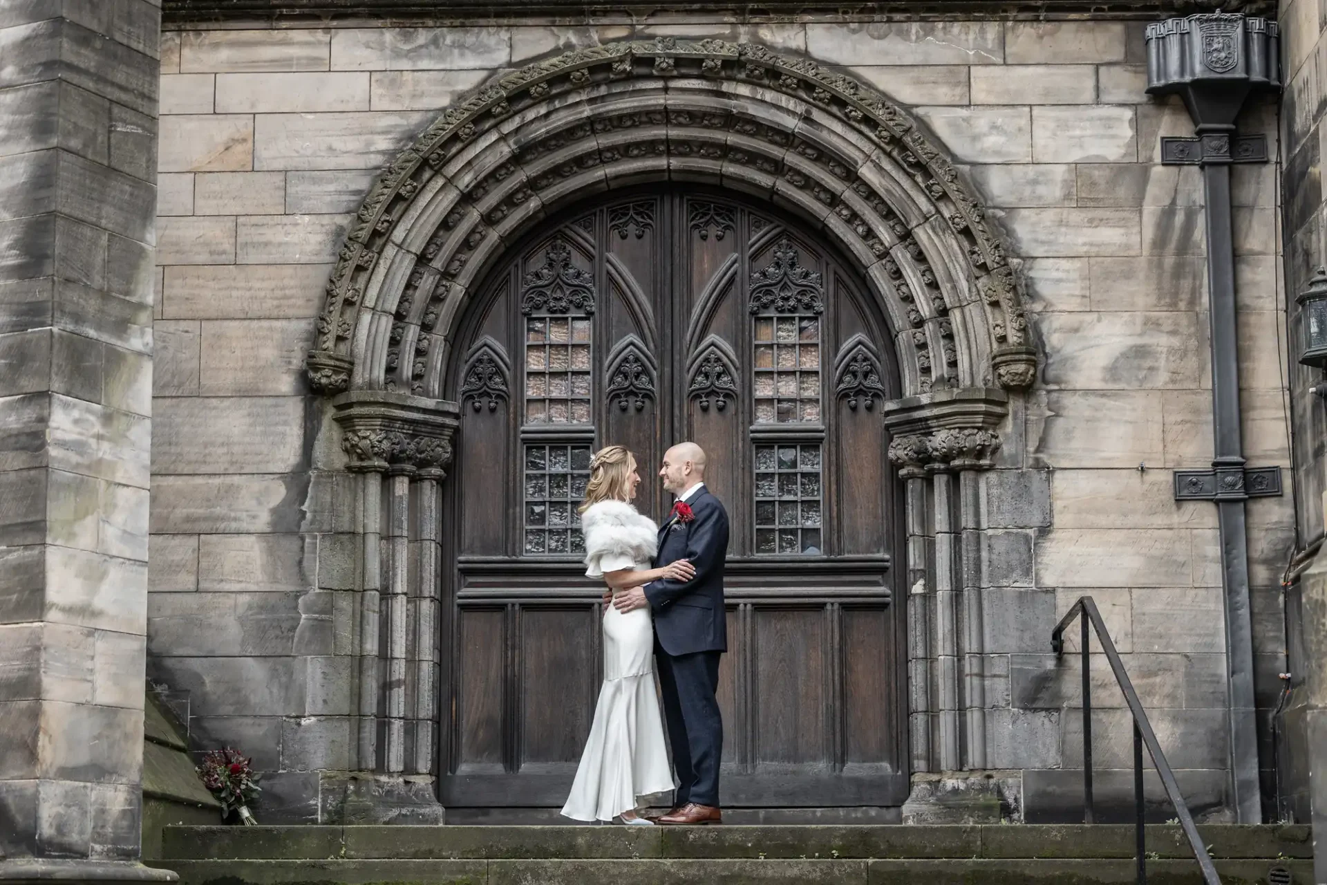 A couple in wedding attire stands embracing in front of an ornate, arched wooden door on a stone building.