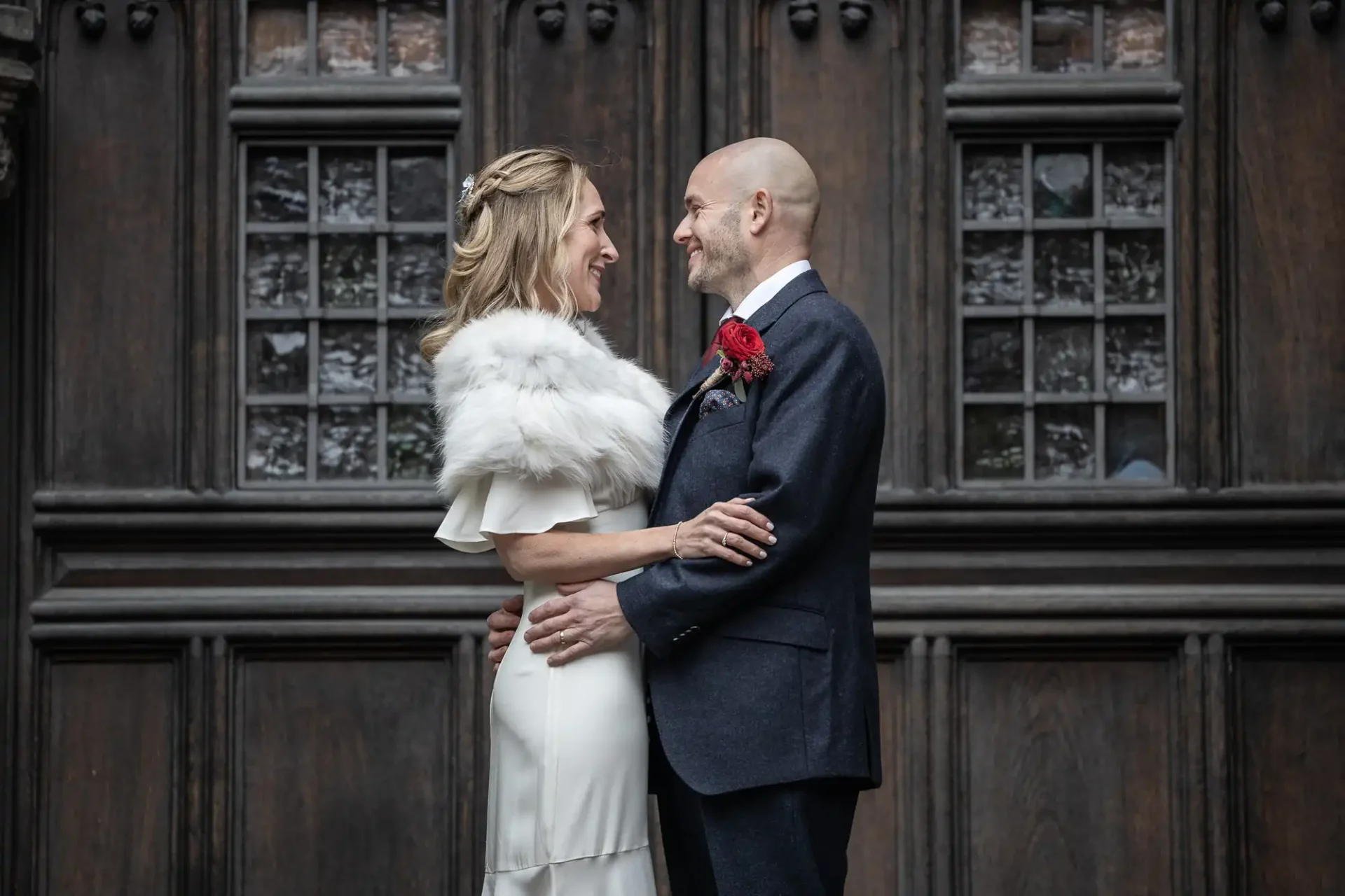 A couple in formal attire warmly embraces, facing each other, in front of a wooden door with glass panels. The woman wears a white dress and fur shawl, while the man is in a dark suit.