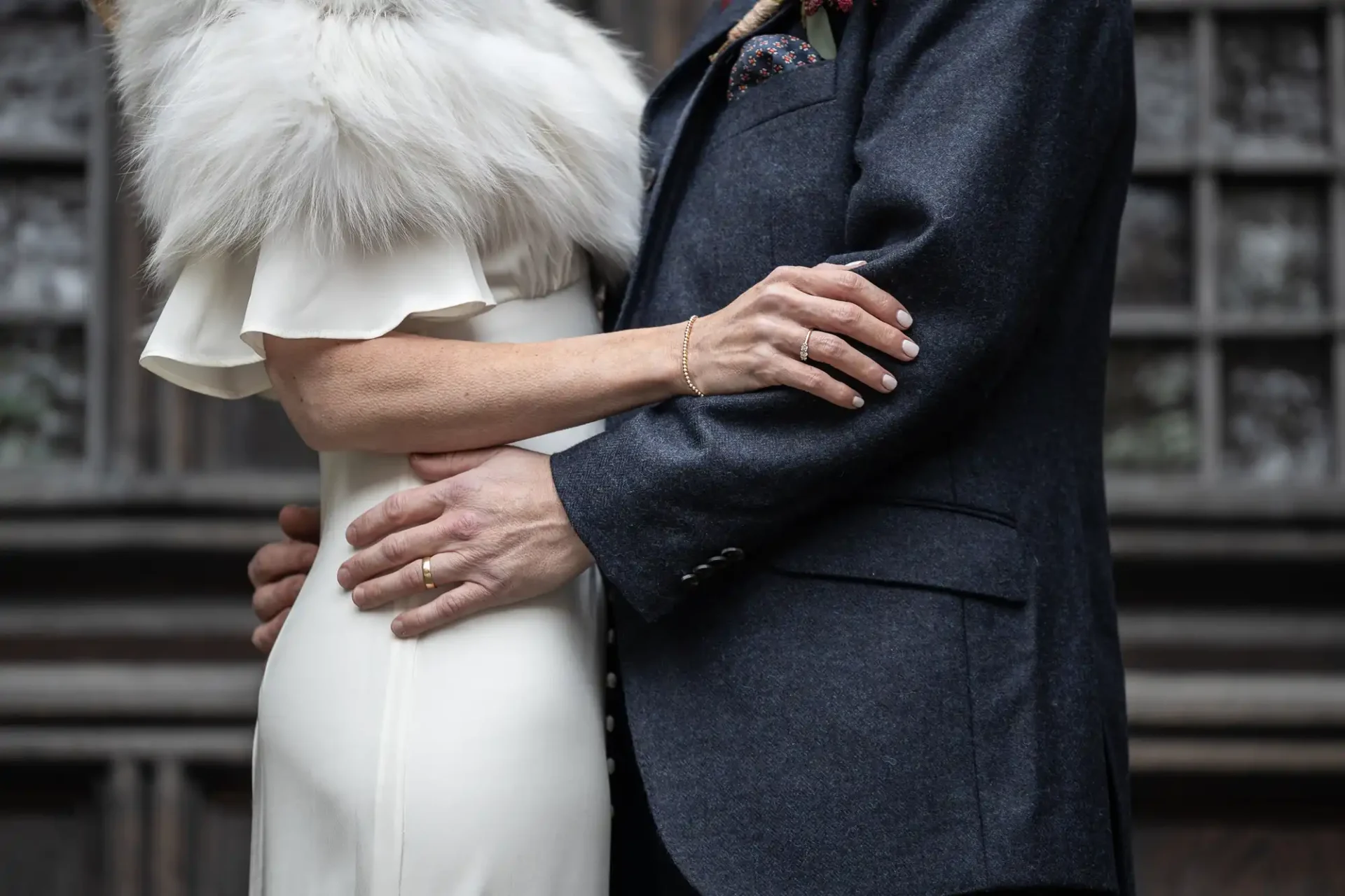 A couple embraces, wearing formal attire. The woman is in a white dress with a fur stole, and the man is in a dark suit. Their wedding bands are visible.