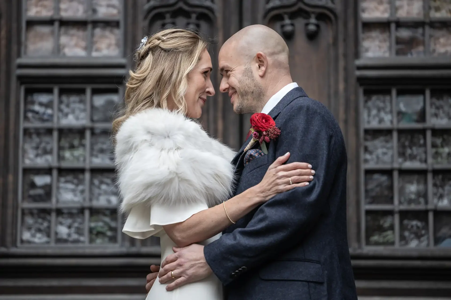 A couple in formal attire embraces in front of ornate windows, gazing at each other. The woman wears a white dress with a fur stole, and the man wears a suit with a red boutonniere.