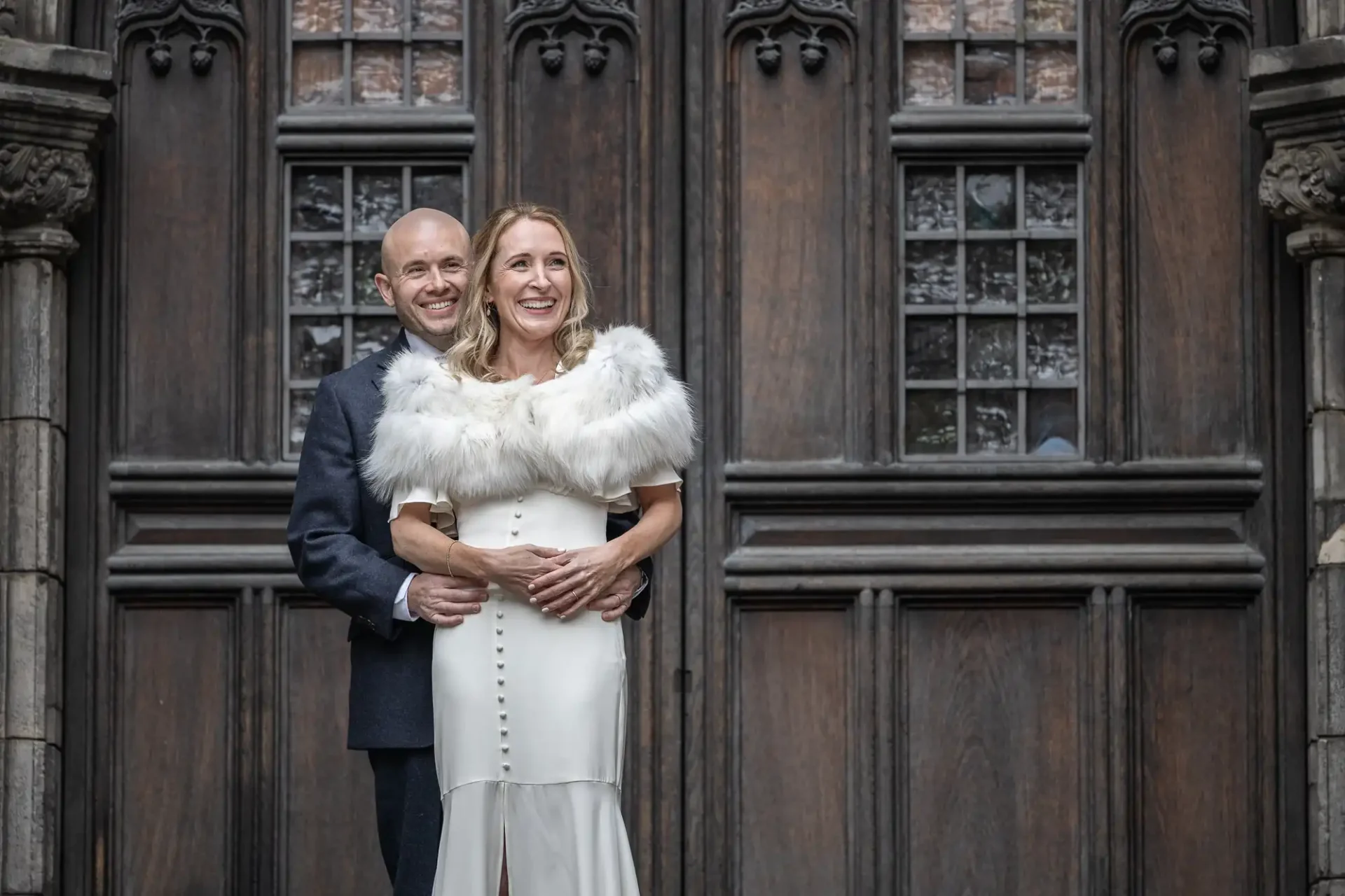 A couple stands in front of a large wooden door, with the man in a dark suit and the woman in a white dress and fur shawl. They smile while embracing and looking forward.