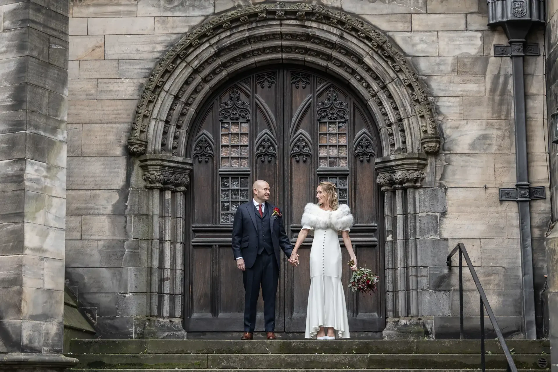 A couple stands on stone steps, holding hands, in front of an ornate arched wooden door. The groom is in a suit, and the bride wears a white dress with a fur stole, holding a bouquet.