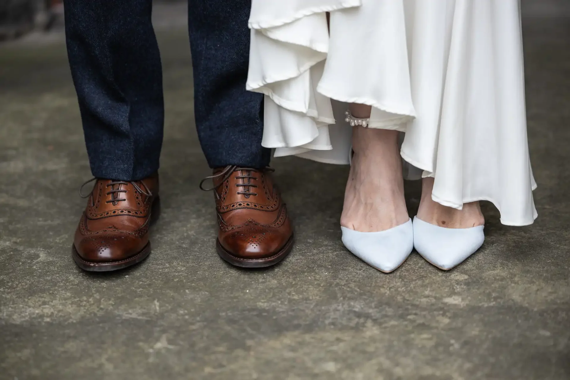 Close-up of a bride and groom's feet. The groom wears brown leather brogues, and the bride wears white pointed heels. Her white dress drapes slightly above the ground.