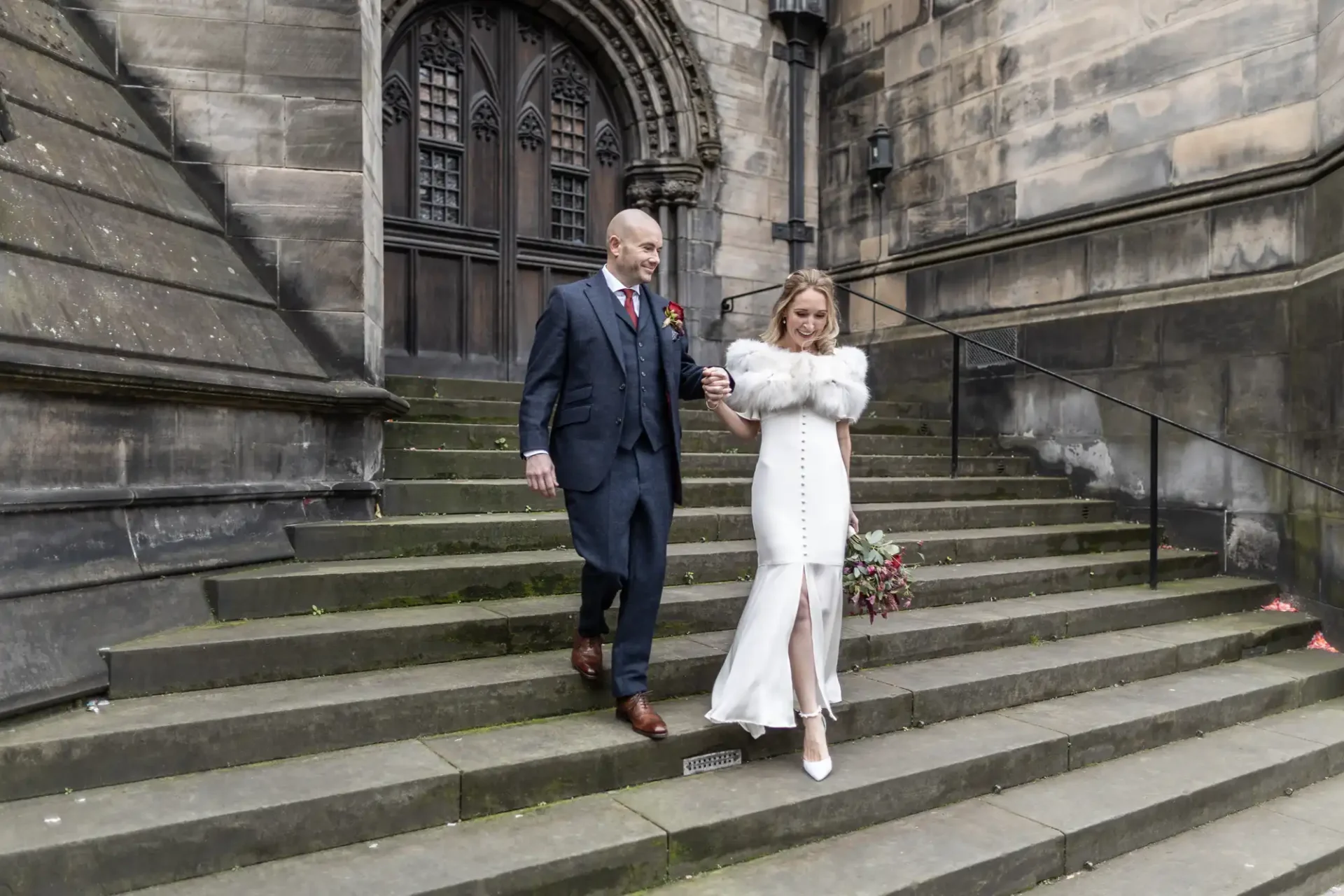 A couple descends stone steps outside a large building. The man wears a suit, and the woman wears a white dress with a fur wrap, holding a bouquet.