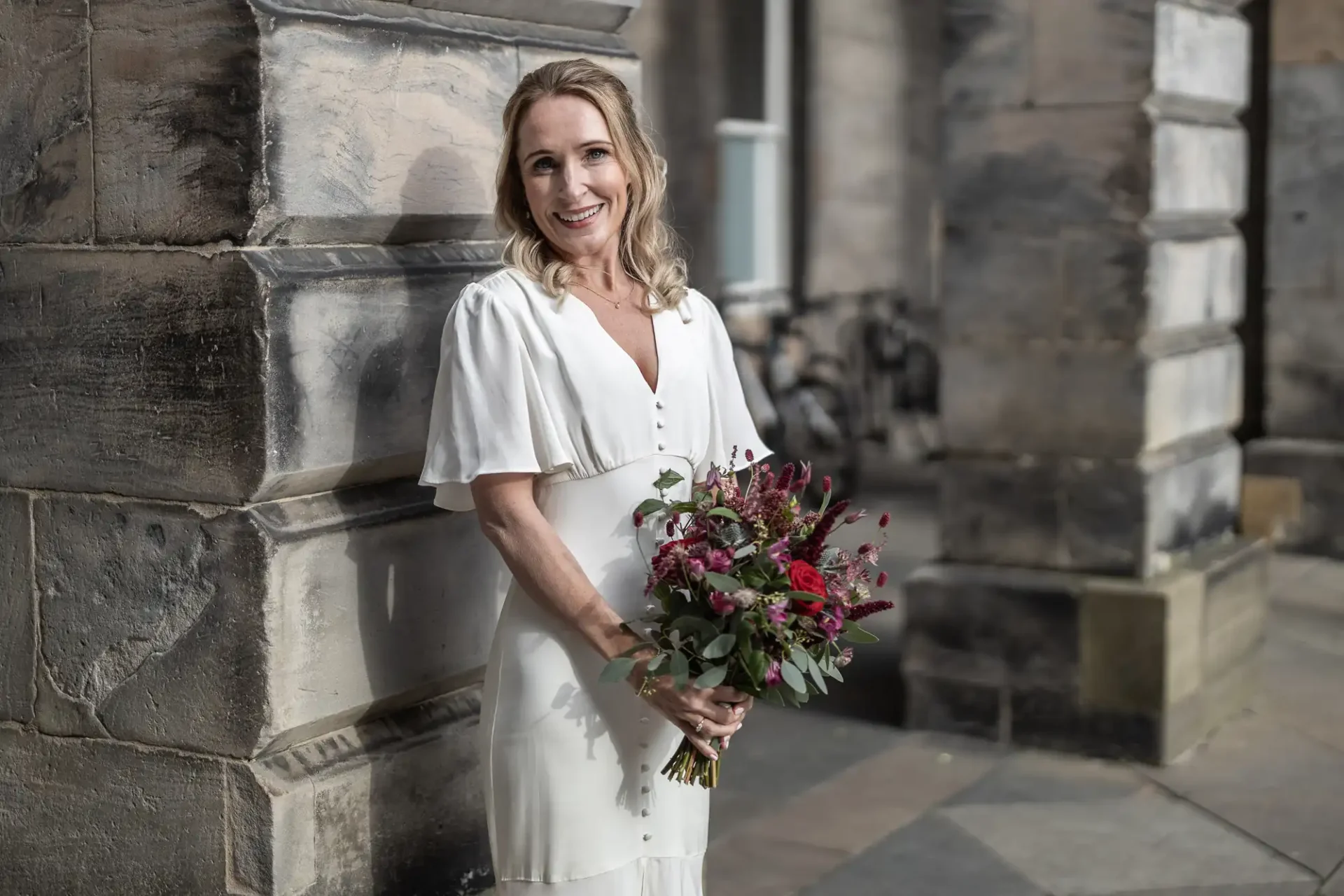 A woman in a white dress holds a bouquet of flowers while standing against a stone wall.