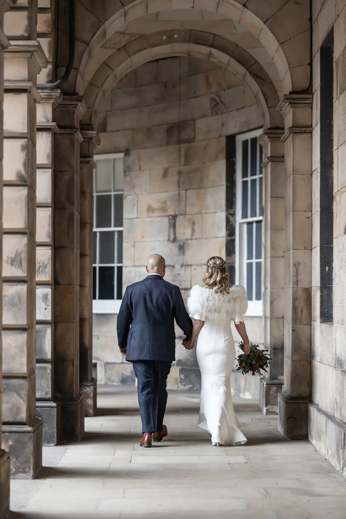 A couple walks hand in hand through an arched stone corridor. The woman wears a white dress and fur shawl, holding a bouquet. The man is in a dark suit.