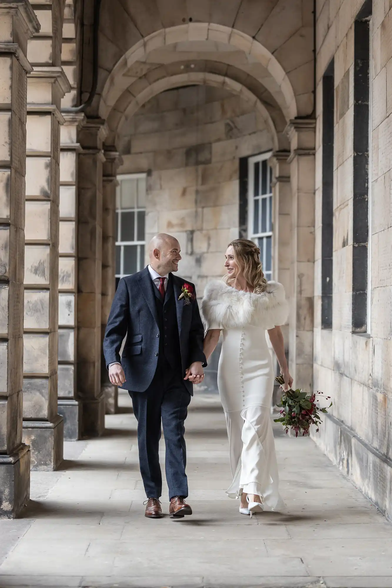 A couple in formal attire walking hand in hand down a stone corridor with archways. She wears a white dress with a fur shawl, holding a bouquet. He wears a dark suit.