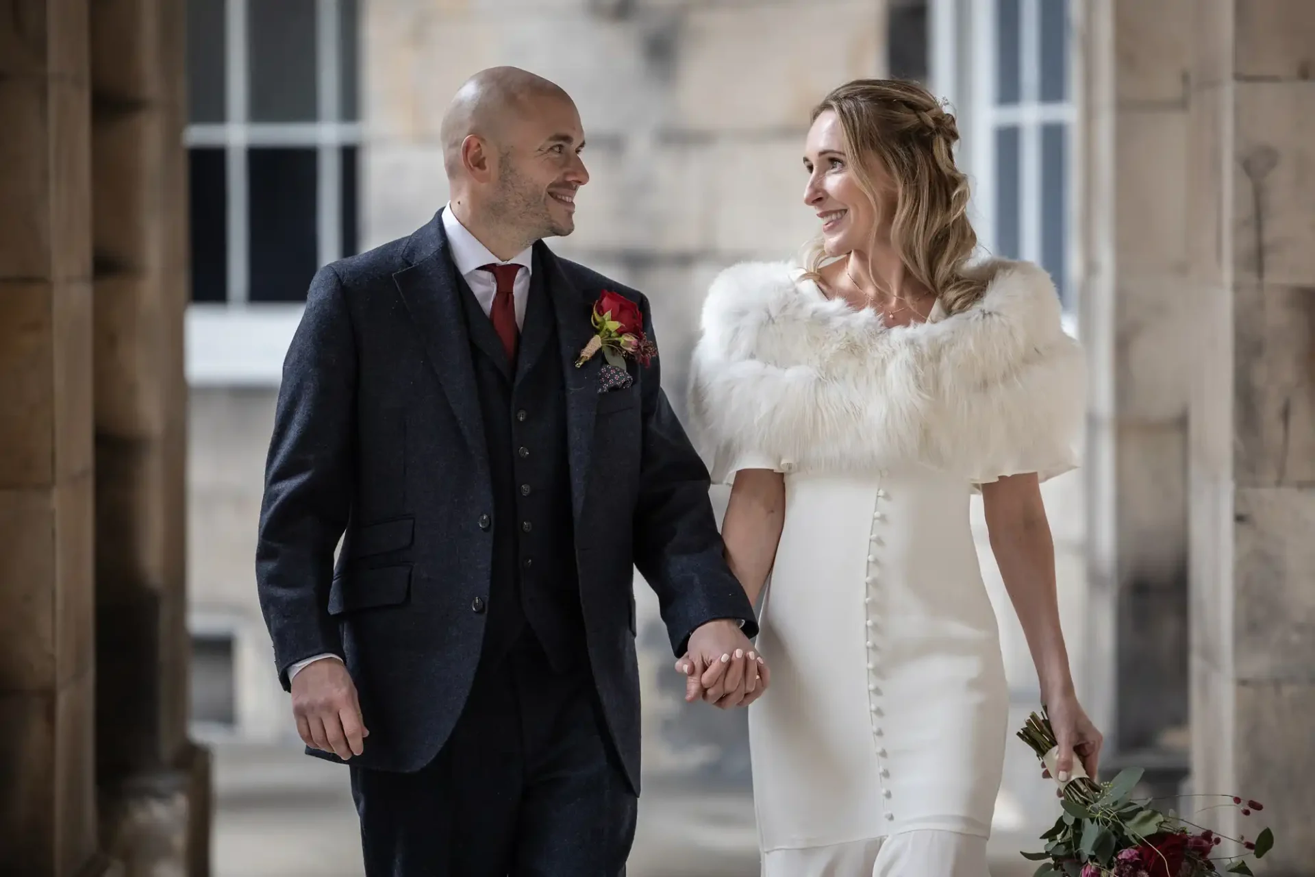 A couple in formal attire walk hand in hand, smiling at each other. The woman wears a white dress with a fur shawl, the man is in a dark suit with a red tie. They are outside a stone building.