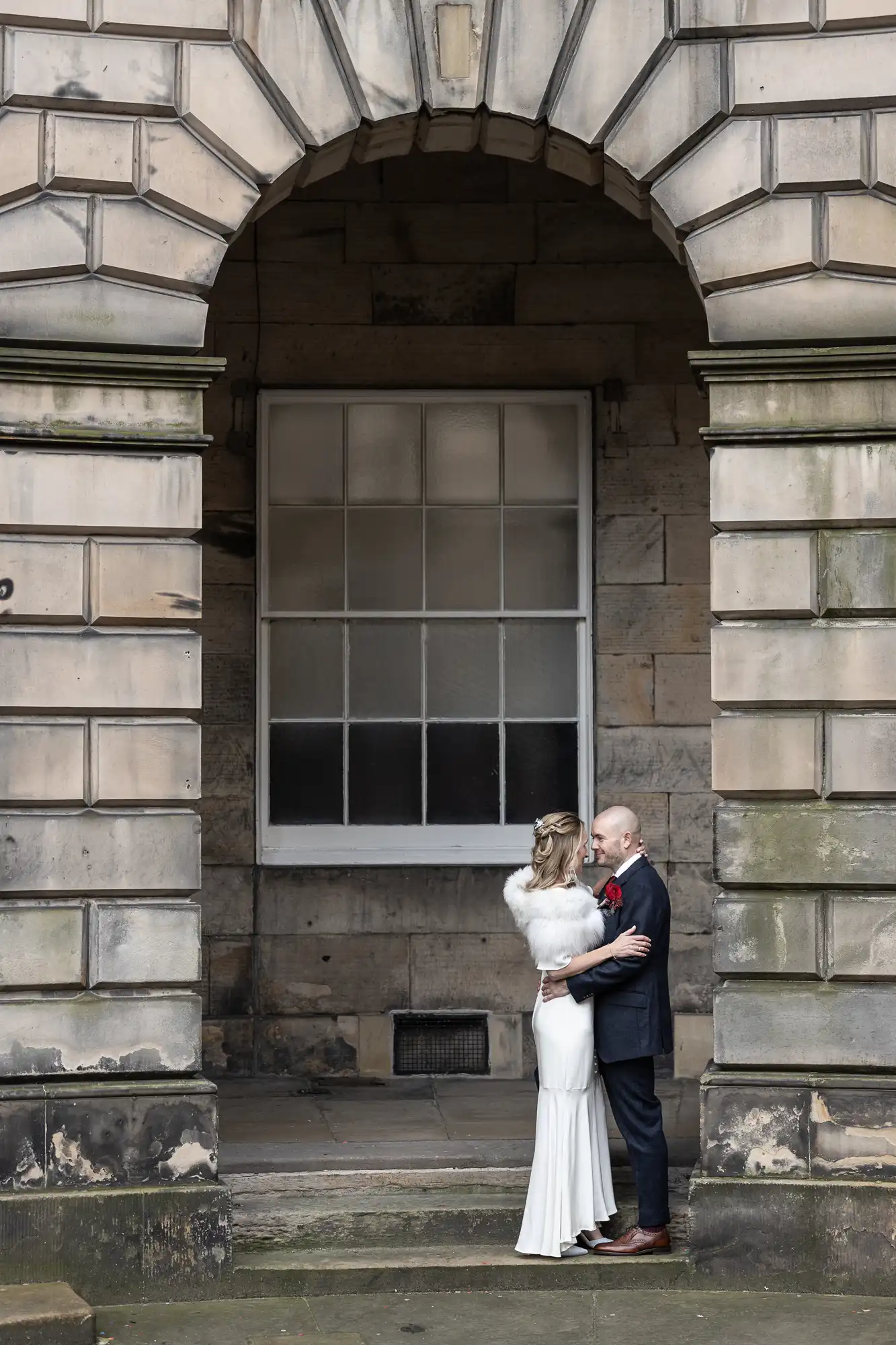 A couple in formal attire embraces under a stone archway with a large window behind them.