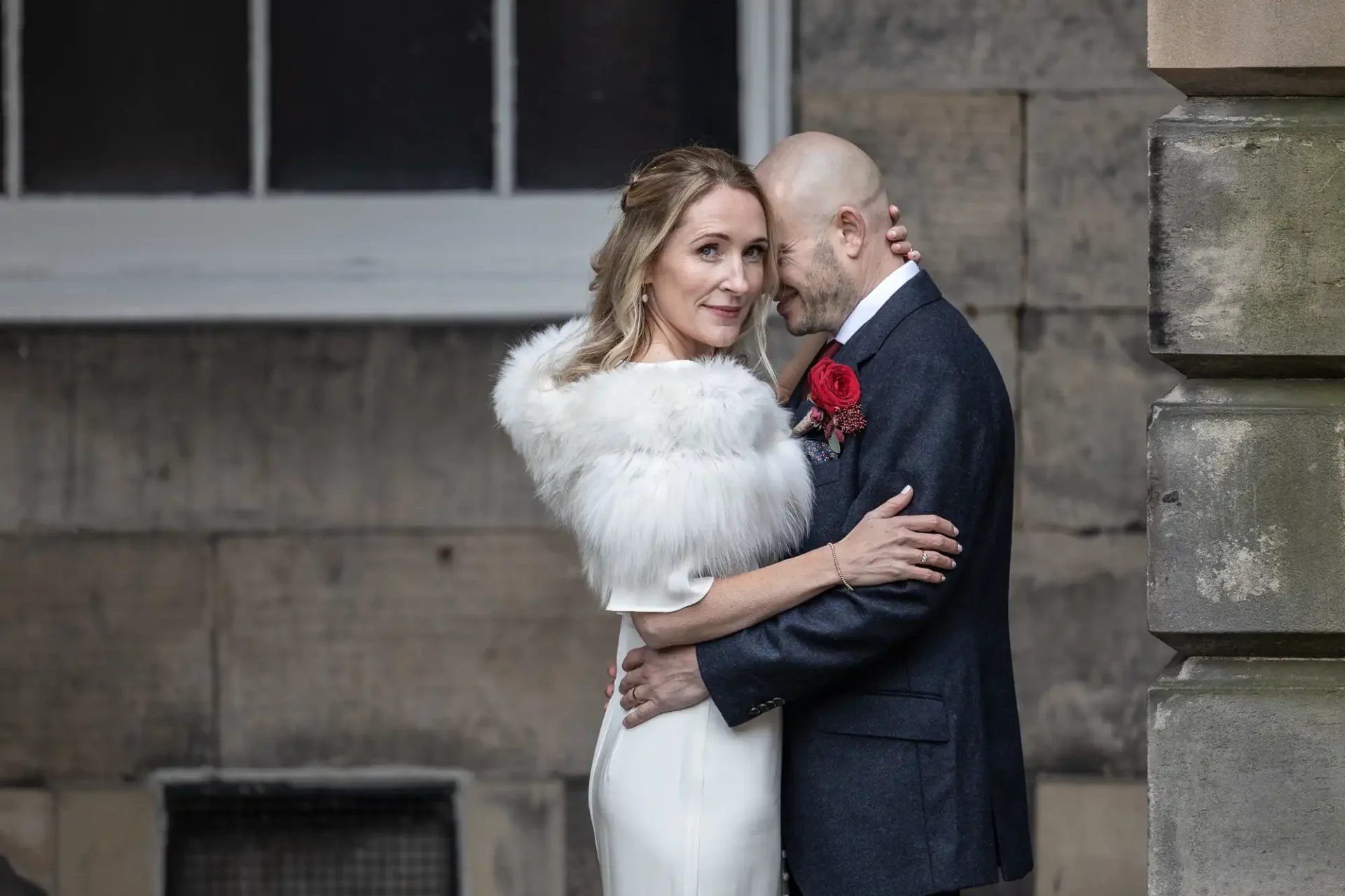 A couple in formal attire, with the woman in a white dress and fur shawl, embracing near a stone building.