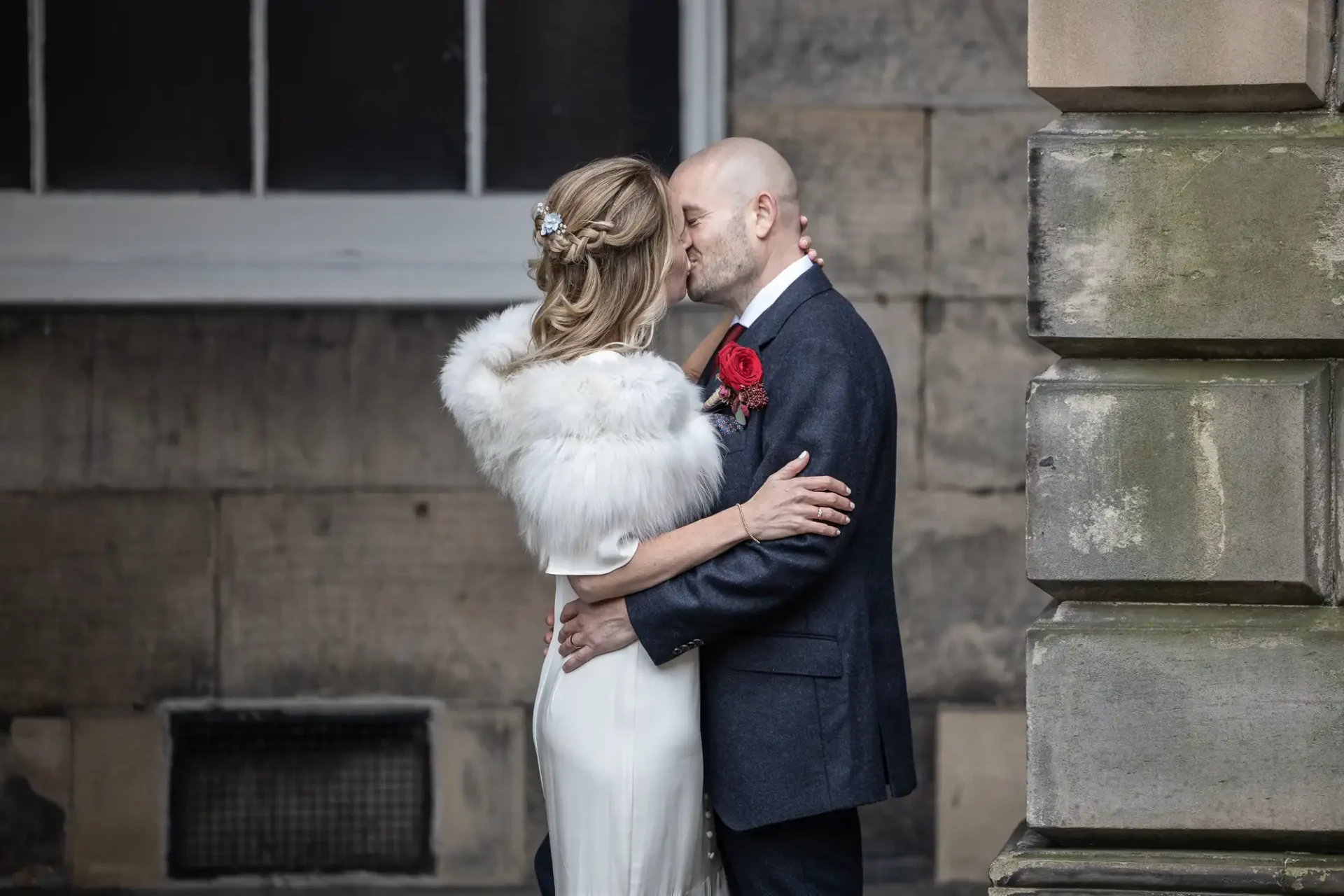 A couple in formal attire shares a kiss outdoors. The woman is wearing a white dress with a fur wrap, and the man is in a dark suit with a red boutonniere.