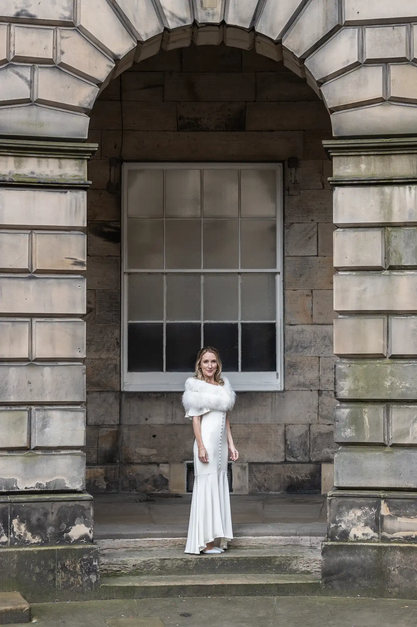 A woman in a white dress and shawl stands under a stone archway with a large window in the background.