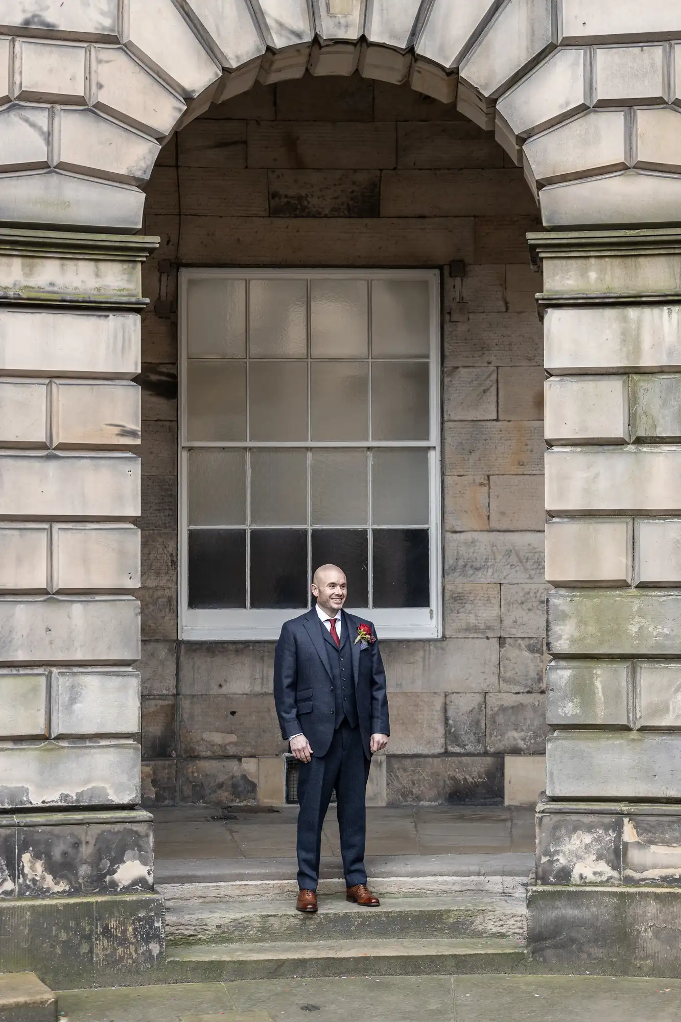 A man in a suit stands under a stone archway in front of a large window, holding a briefcase.