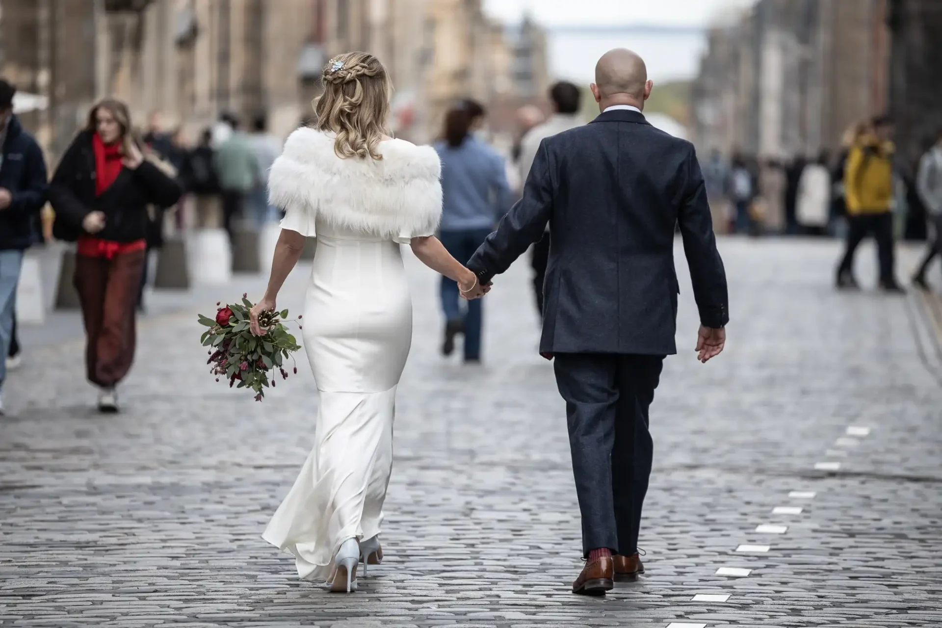 A couple in formal attire holds hands while walking down a cobblestone street, surrounded by people. The woman wears a white dress and fur stole, holding a bouquet.