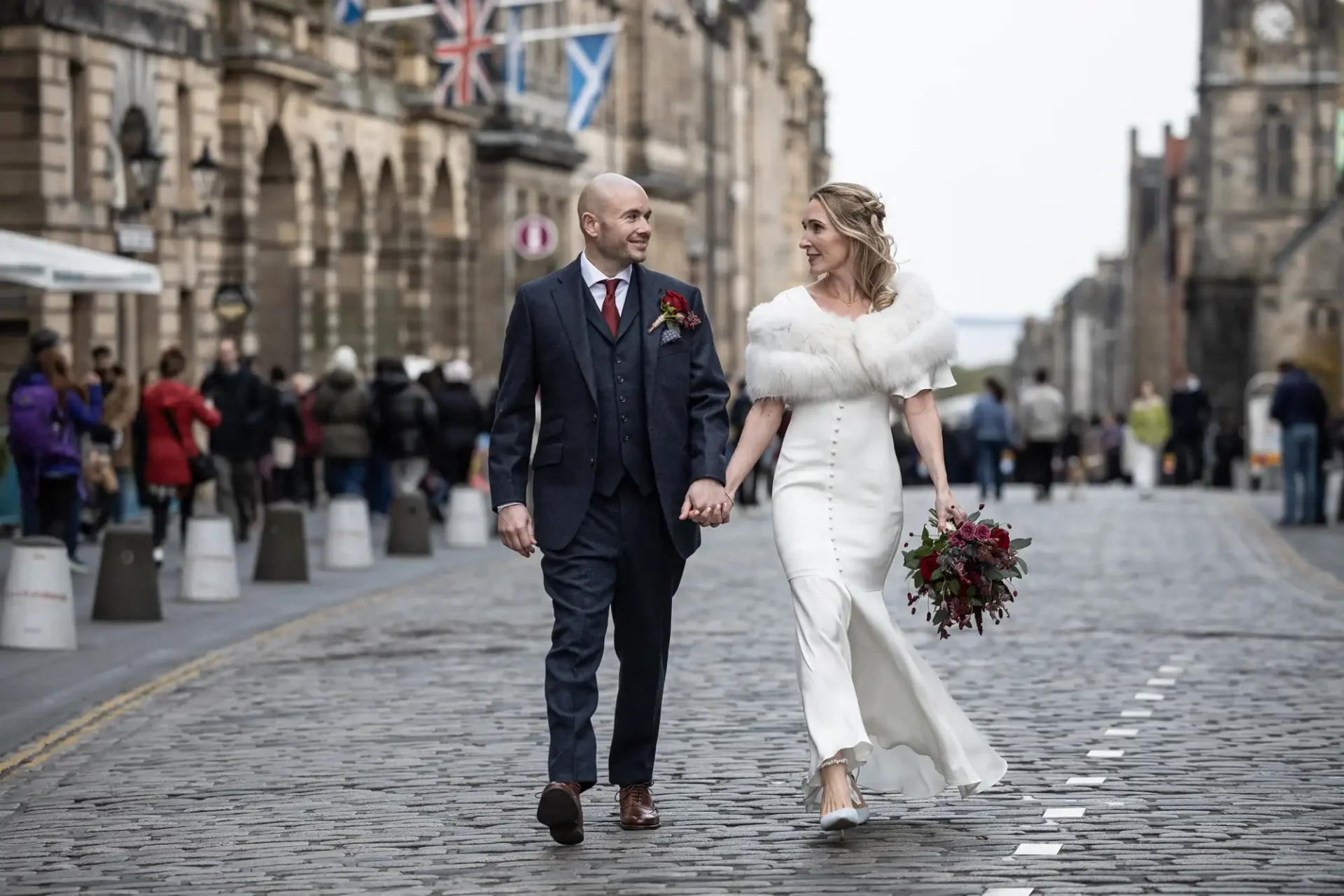 A couple in formal attire walks hand in hand on a cobblestone street. The woman holds a bouquet, and there are flags and people in the background.