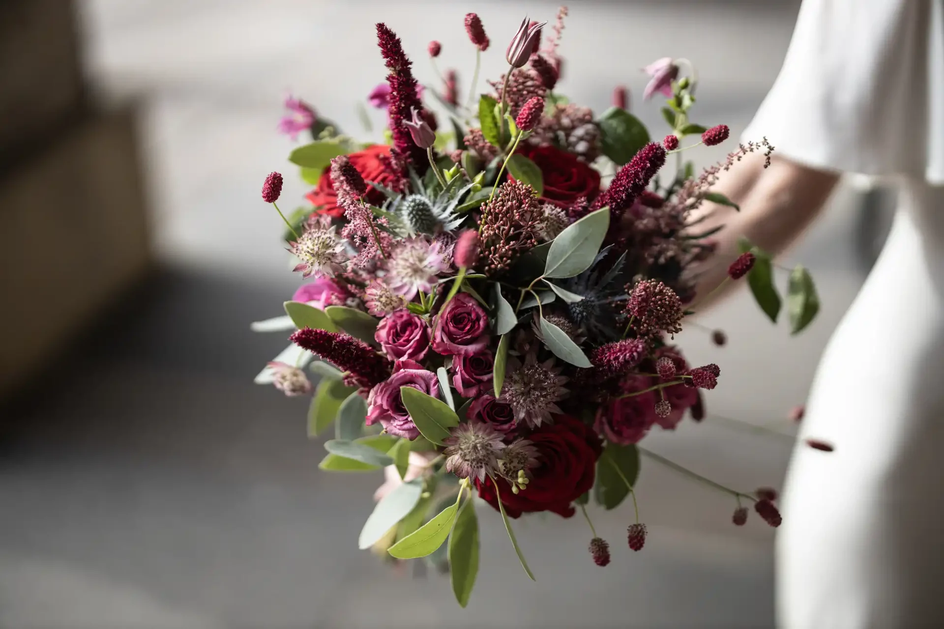 A bouquet with red and pink flowers, green leaves, and various foliage, held by a person in a white outfit.