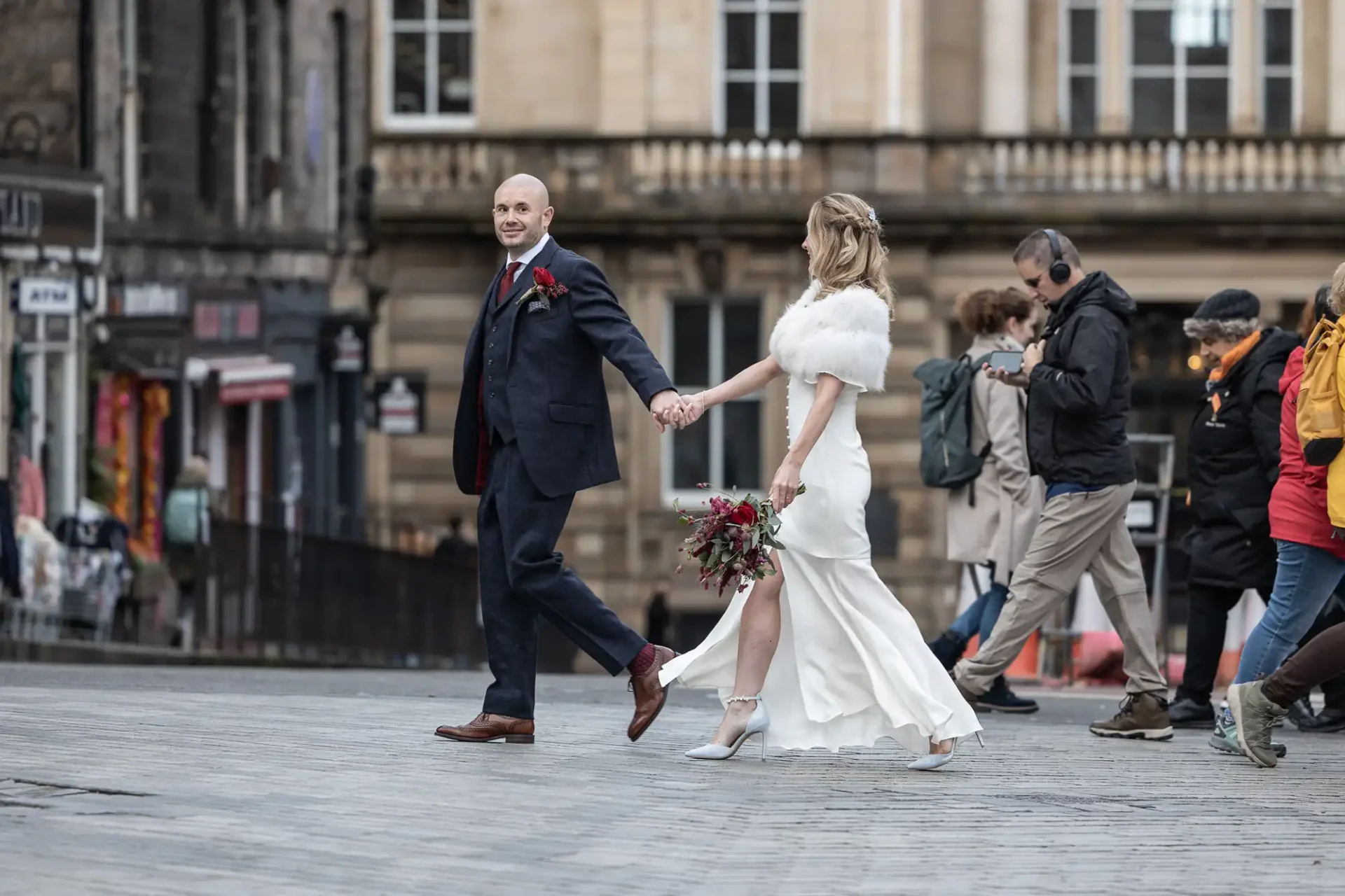 A couple in wedding attire holding hands walks across a city street. The groom wears a suit and the bride wears a white dress. Pedestrians pass by in the background.