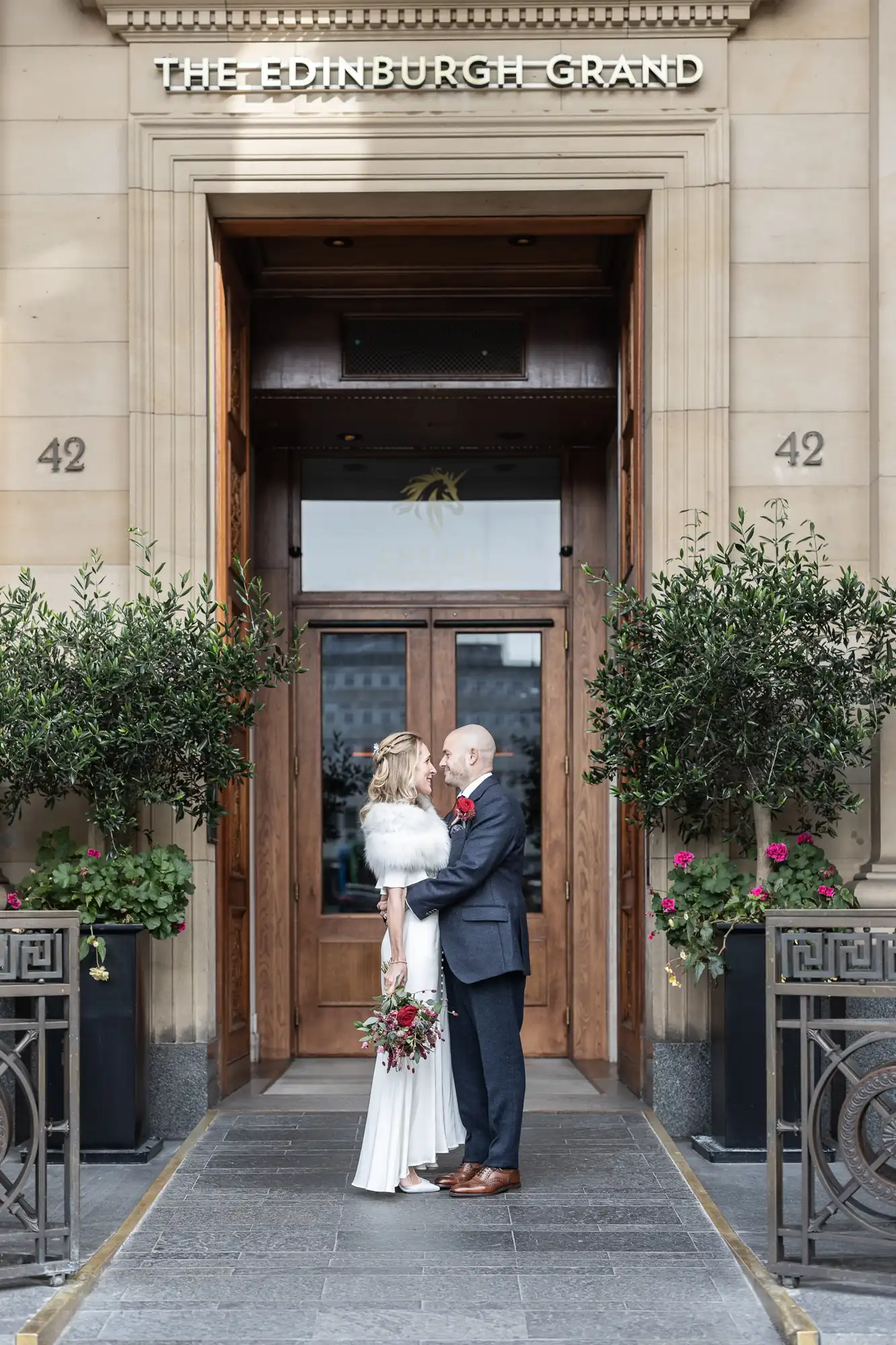 A couple stands embracing outside The Edinburgh Grand building entrance, each holding a bouquet of flowers.