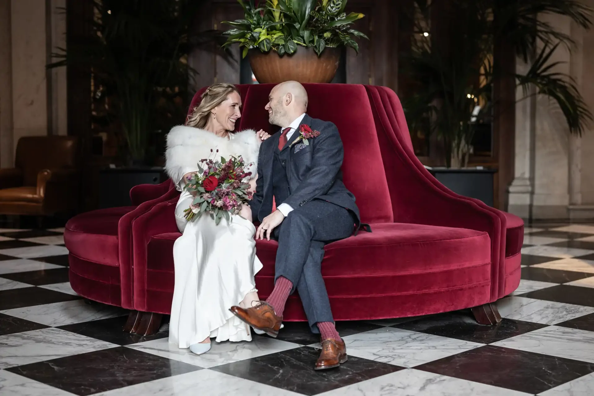 Cheval The Edinburgh Grand wedding photographer image of a bride and groom sit on a red velvet couch, smiling at each other. The bride holds a bouquet of red and pink flowers. The setting is a stylish room with a checkered floor.