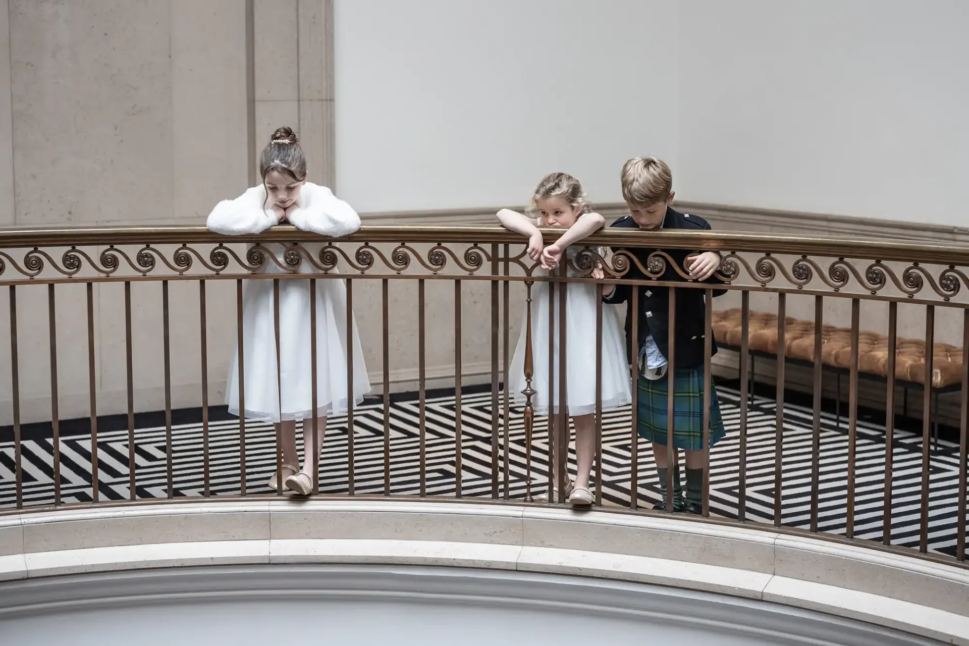 Three children in formal attire lean on a railing, looking down. The floor below features a black and white geometric pattern.