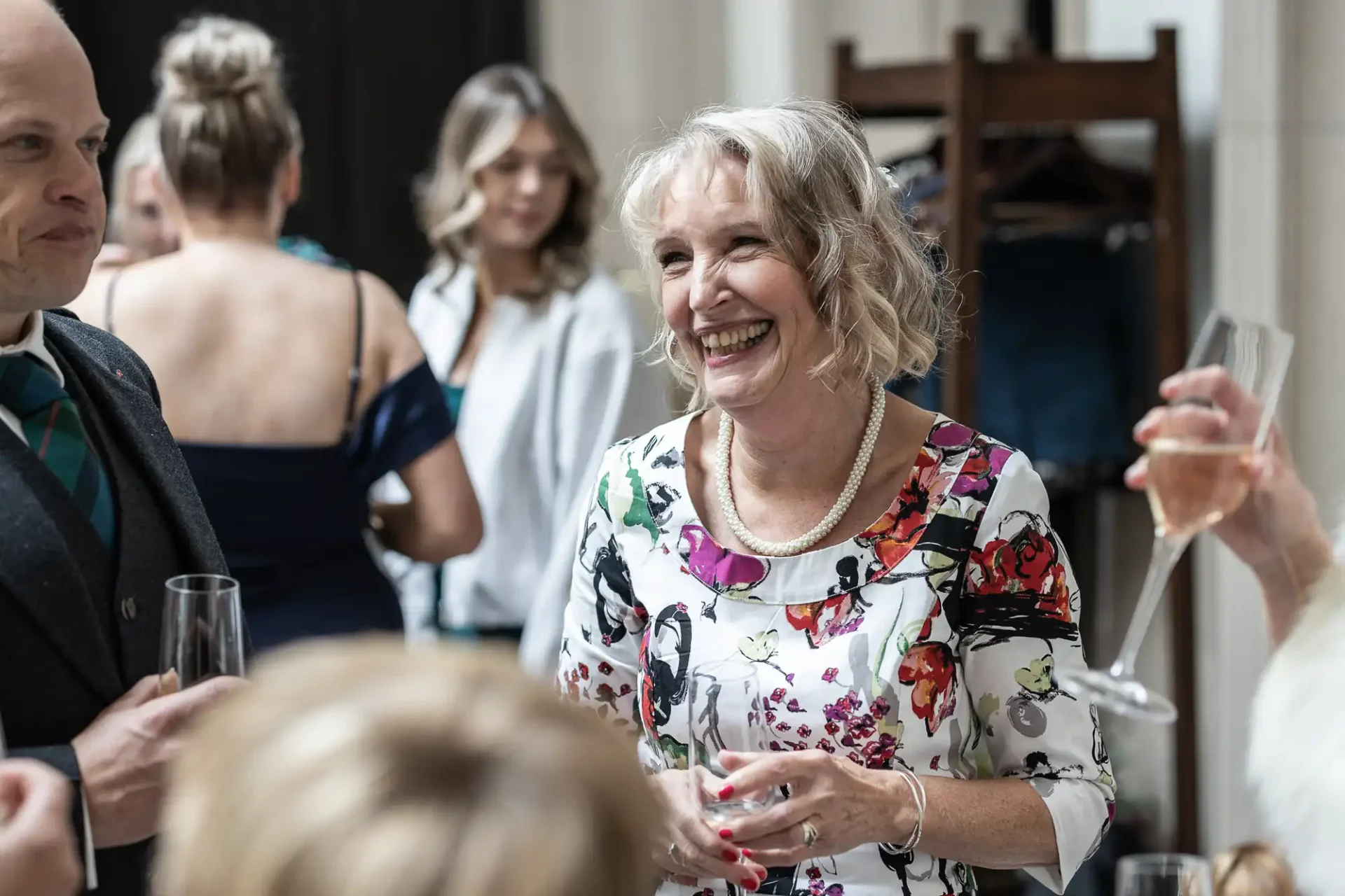 A woman in a floral dress smiles and holds a glass at a social gathering, surrounded by other people.