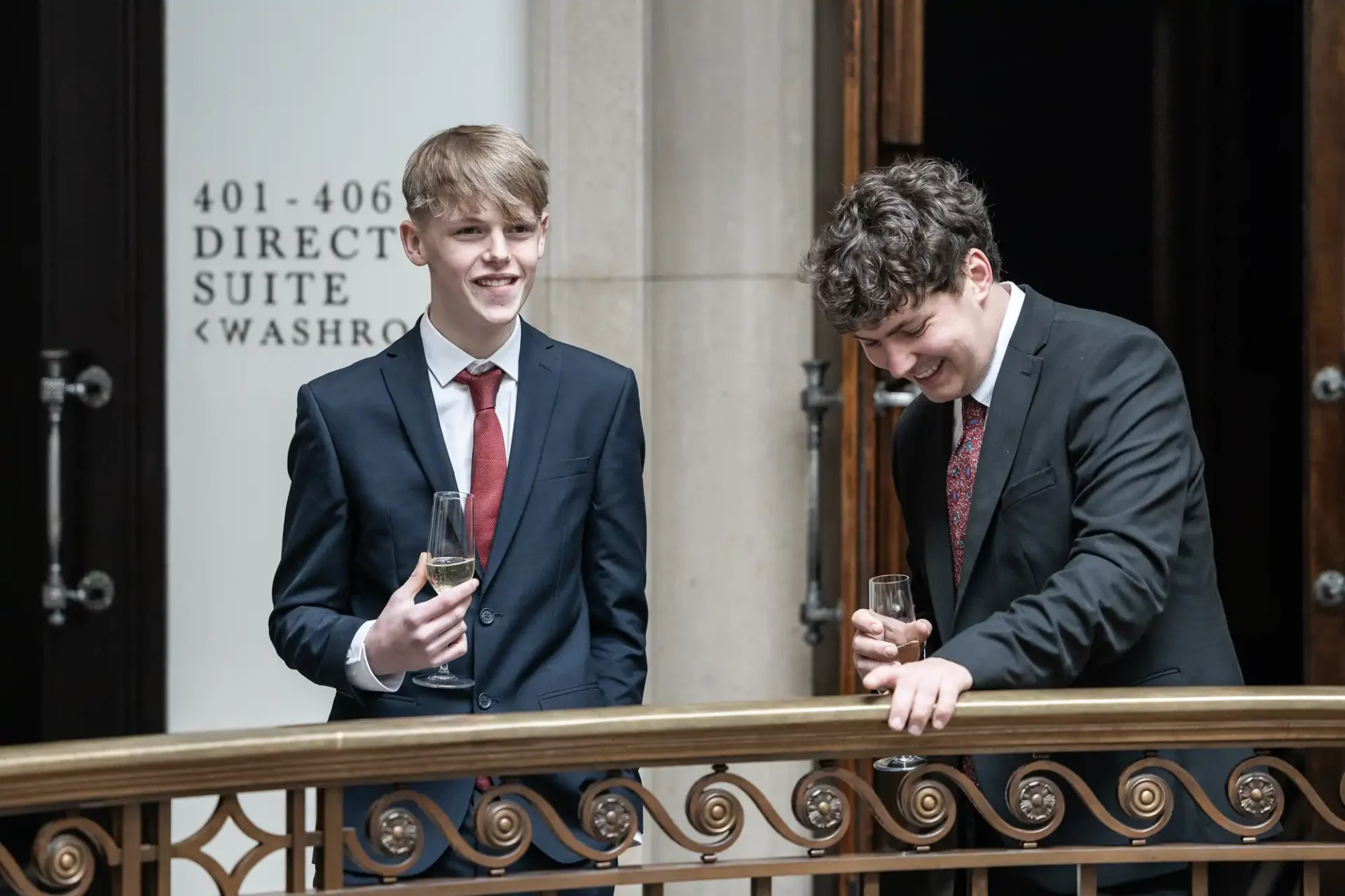 Two young men in suits stand by a railing, holding glasses. One is smiling and looking away; the other is looking down, also smiling. A sign indicating directions is visible on the wall behind them.