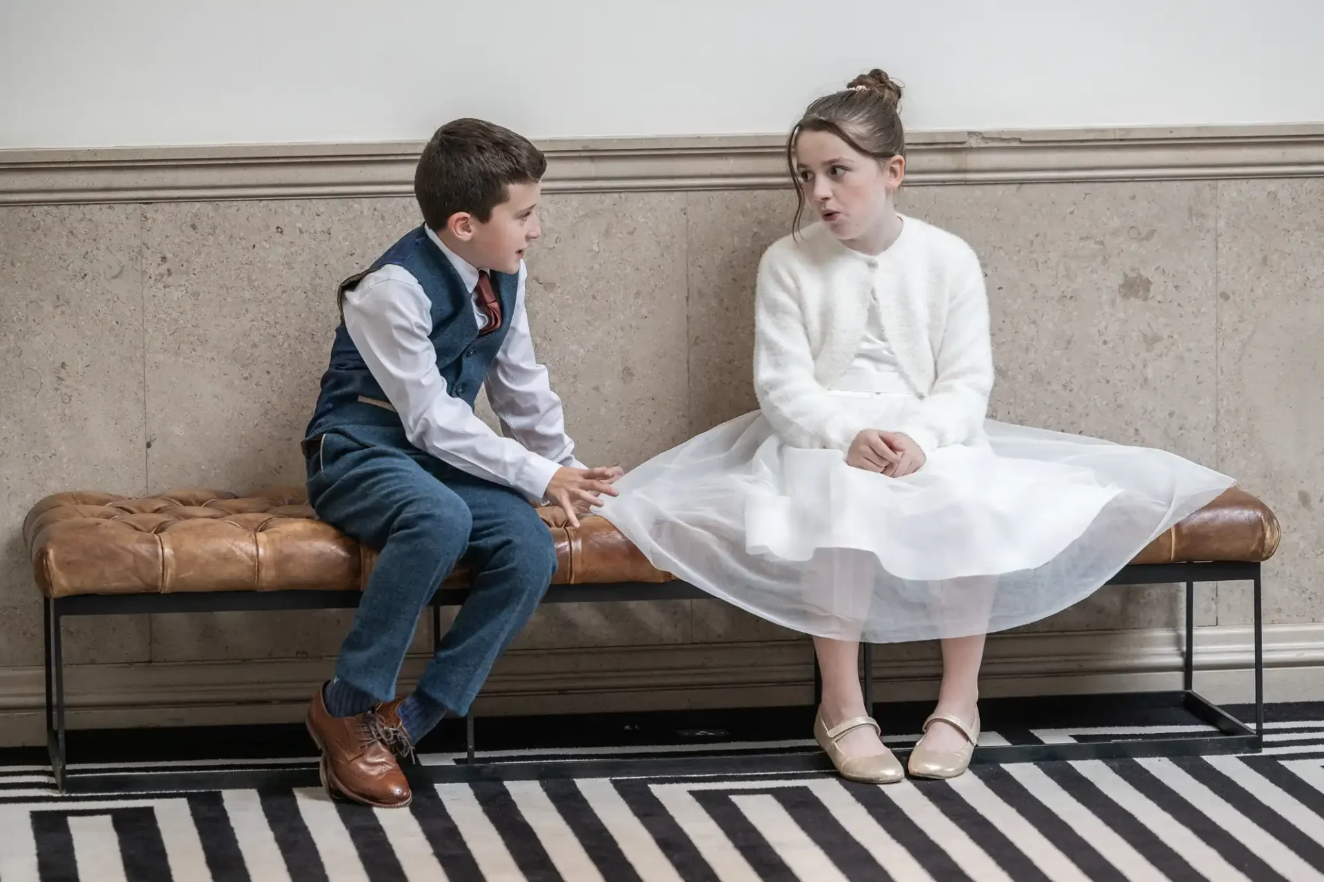 A young boy and girl sit on a bench indoors. The boy is wearing a suit, and the girl is in a white dress and cardigan. They are looking at each other while seated.