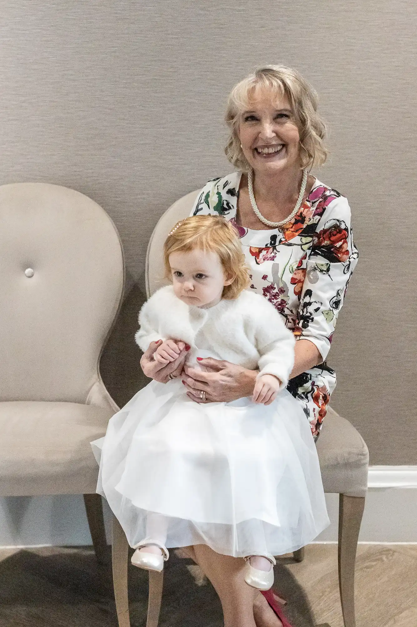 Woman in a floral dress sits on a chair holding a young child in a white dress and cardigan. Both have blonde hair. The child looks away while the woman smiles at the camera.