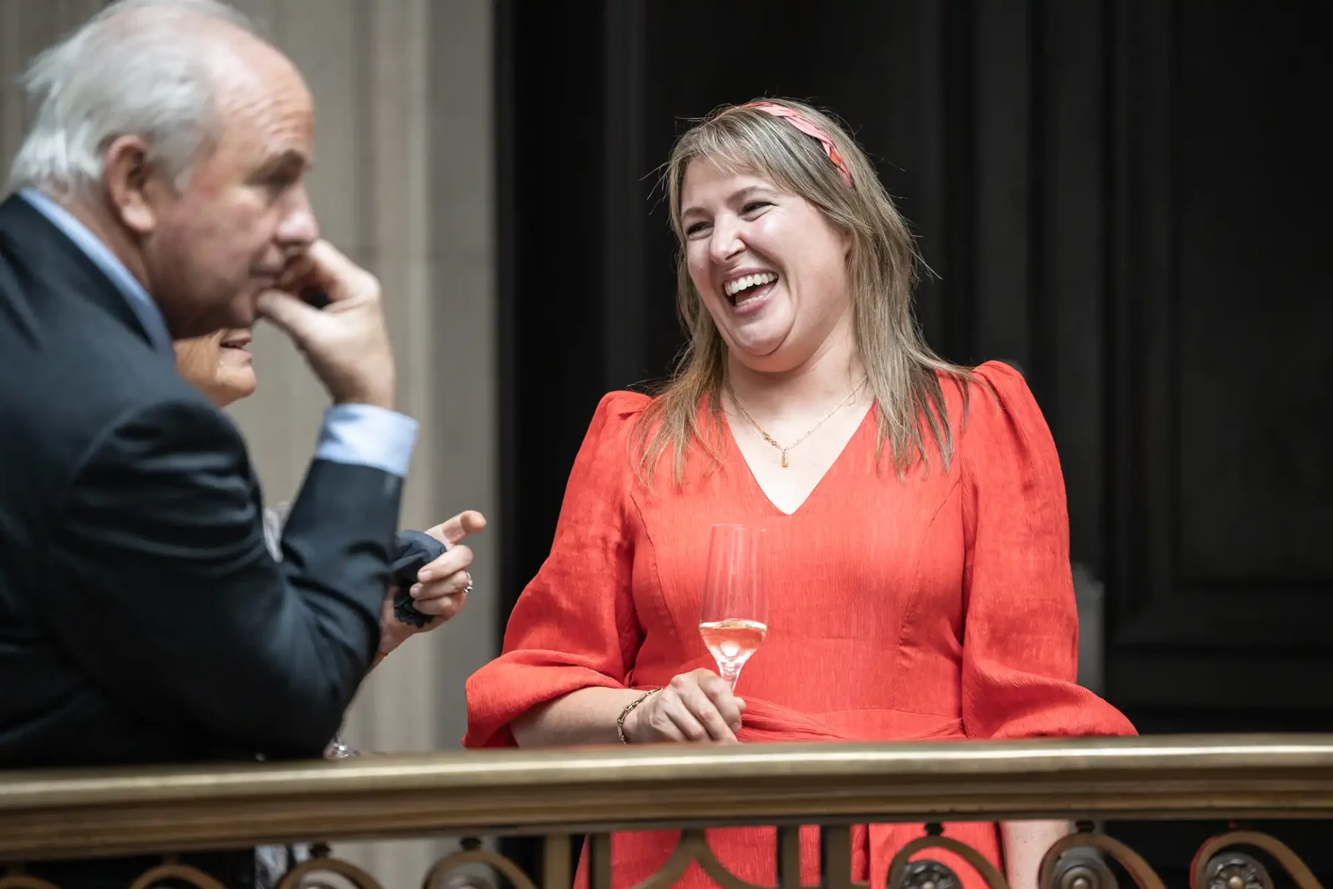 A woman in a red dress smiles while holding a glass. Two people are beside her, one in focus and one partly obscured. They are standing near a railing indoors.