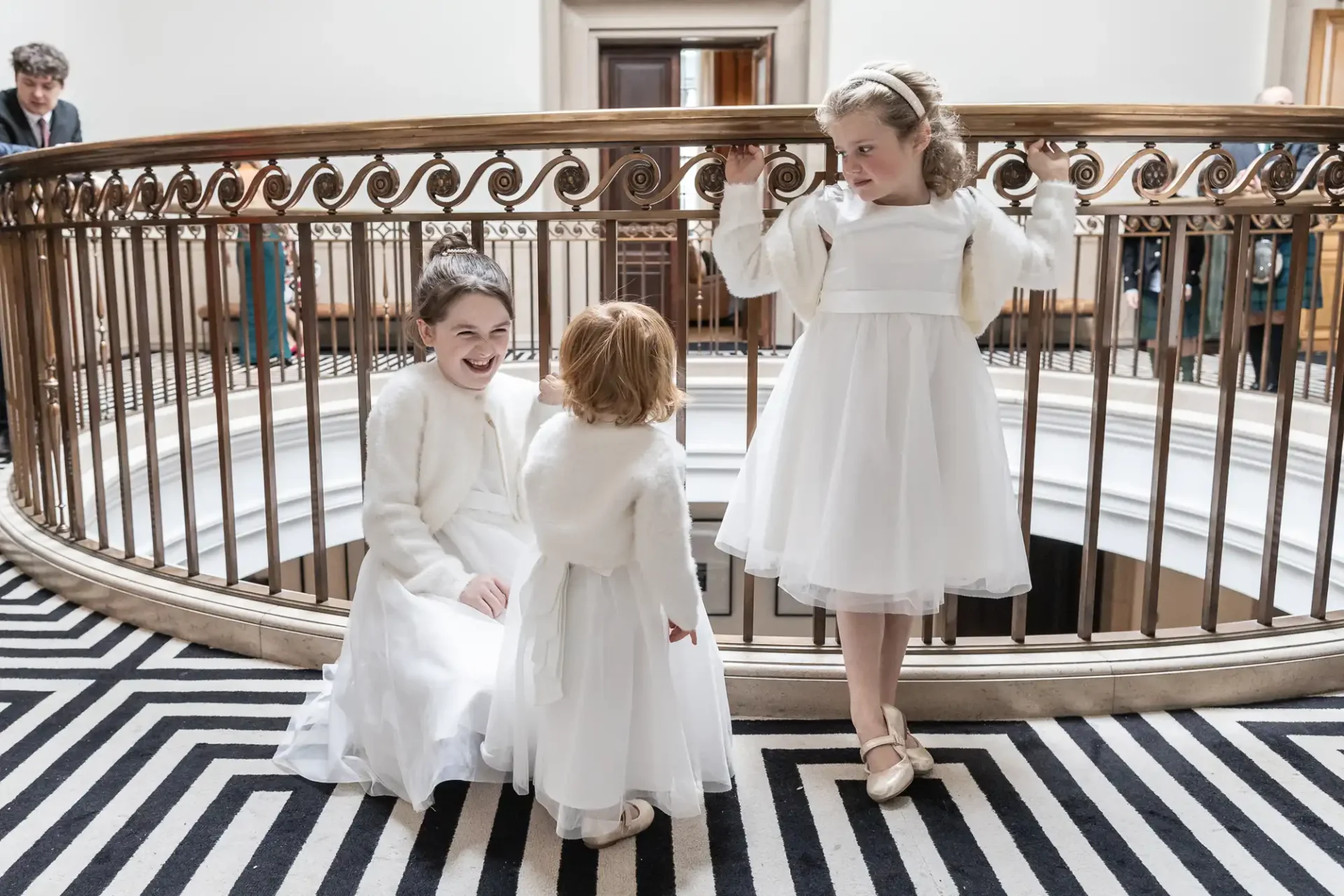 Three young girls in white dresses stand by an ornate railing. Two are smiling, one with hands on the rail. A person in the background. Black-and-white zigzag carpet below.