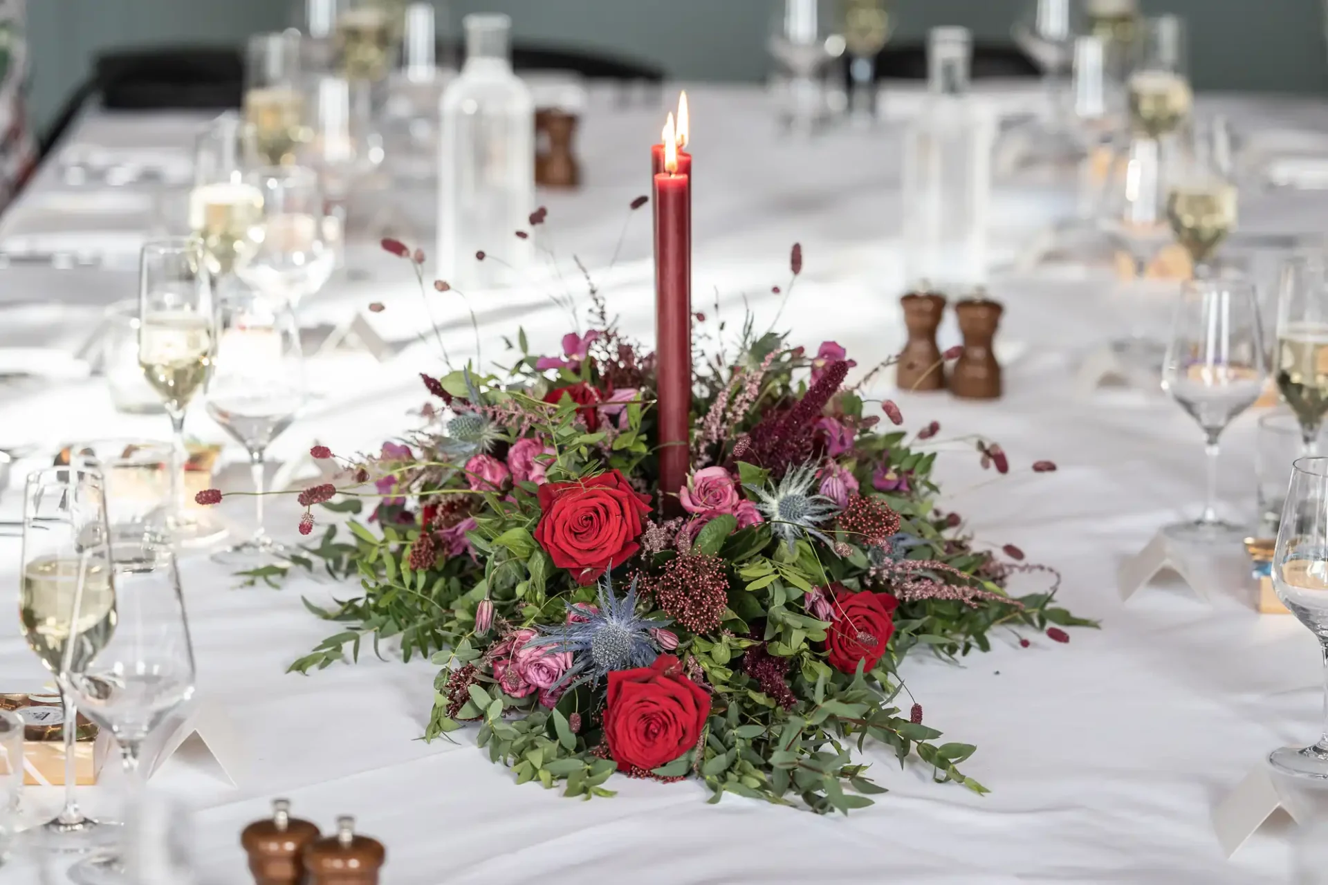 Floral centerpiece with red roses and greenery, featuring a single tall lit red candle. Surrounded by wine glasses and bottles on a white tablecloth.