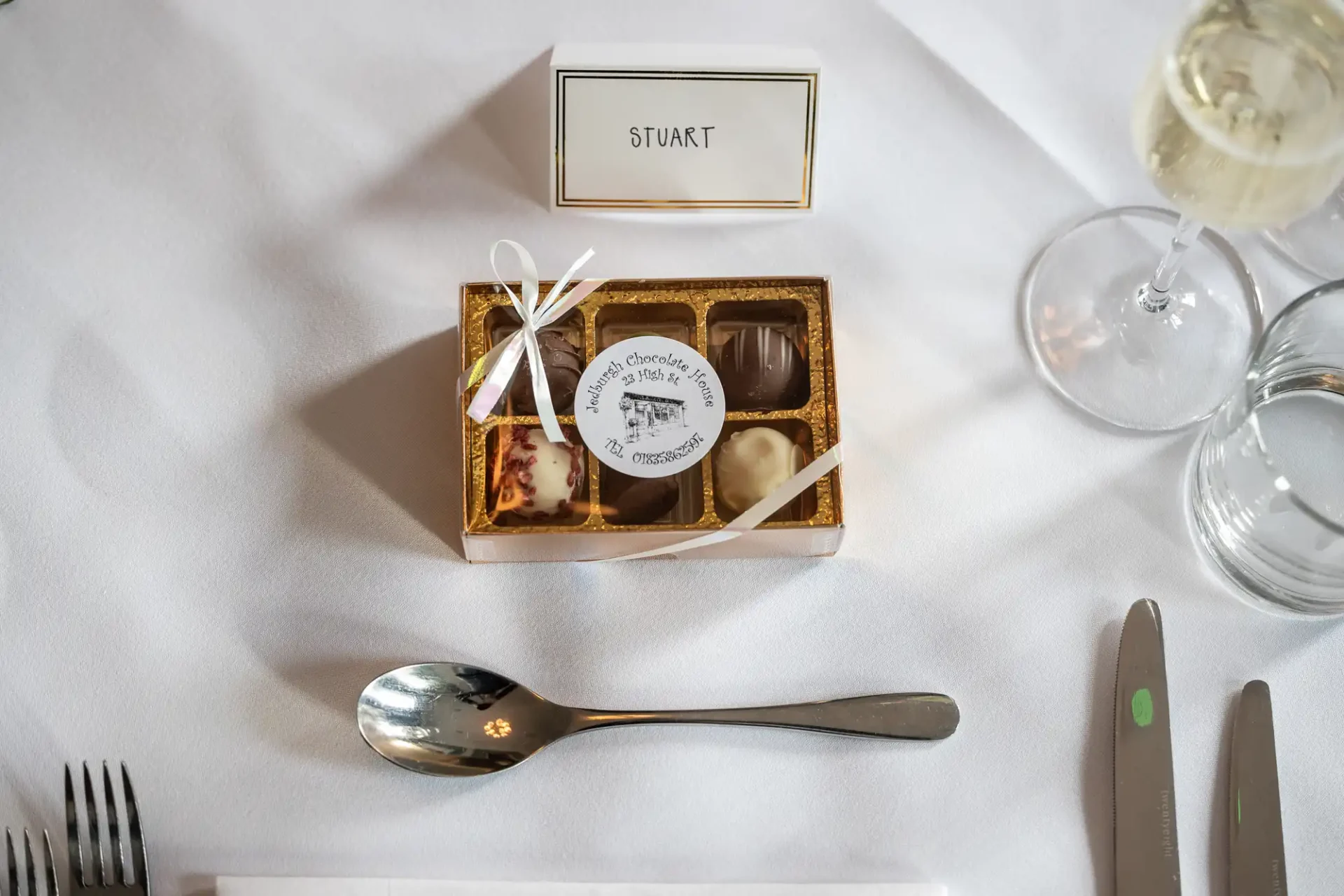 A small box of assorted chocolates with a ribbon sits on a white tablecloth next to a place card labeled "Stuart," a spoon, knife, fork, glass of water, and a glass of white wine.