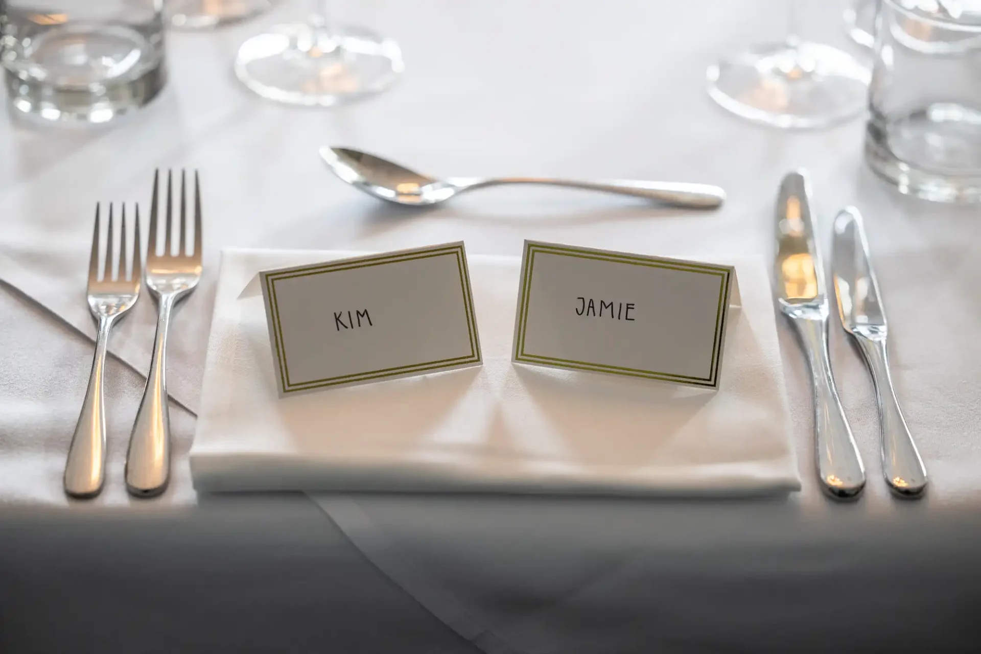 Table setting with two name cards labeled "Kim" and "Jamie" on a white napkin, surrounded by cutlery and glasses.