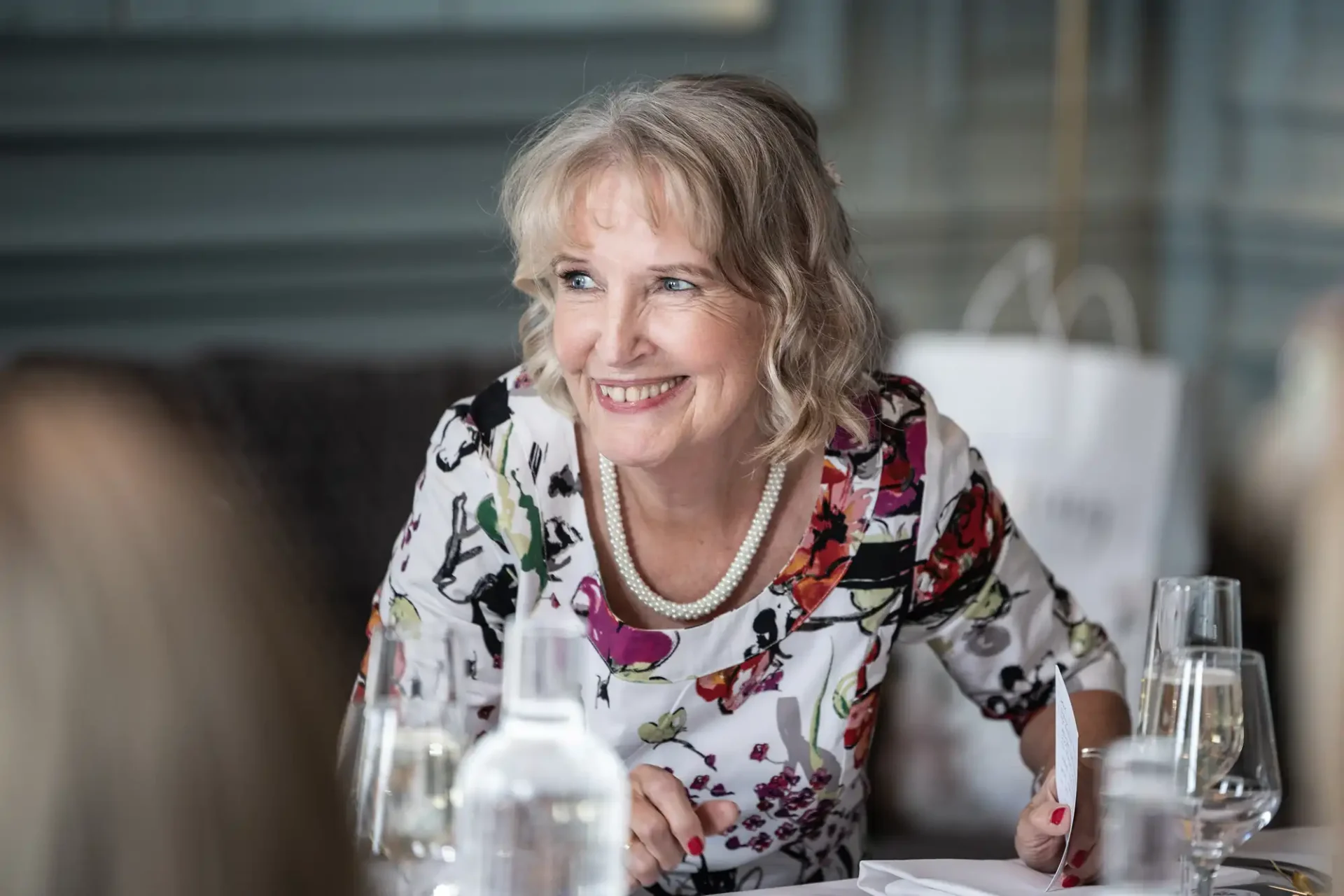 A woman with shoulder-length blonde hair smiles while sitting at a table with dishes and glasses. She wears a floral top and a pearl necklace.