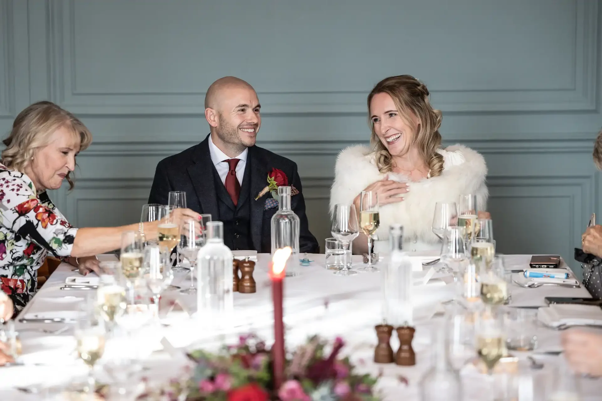 A man and a woman in formal wear smile at a dining table with others. Glasses, bottles, and a floral centerpiece are on the table.