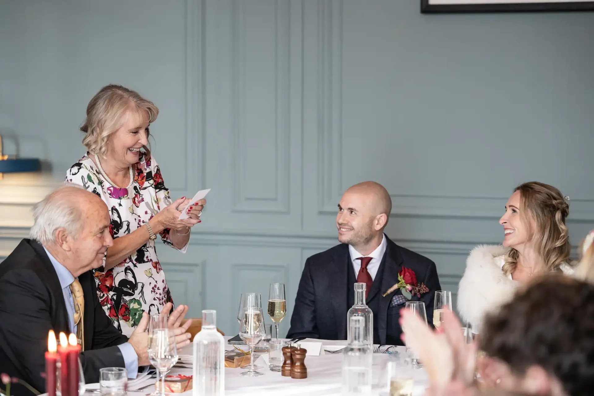 A woman gives a speech at a formal table setting with three seated individuals. They appear to be engaged and smiling.