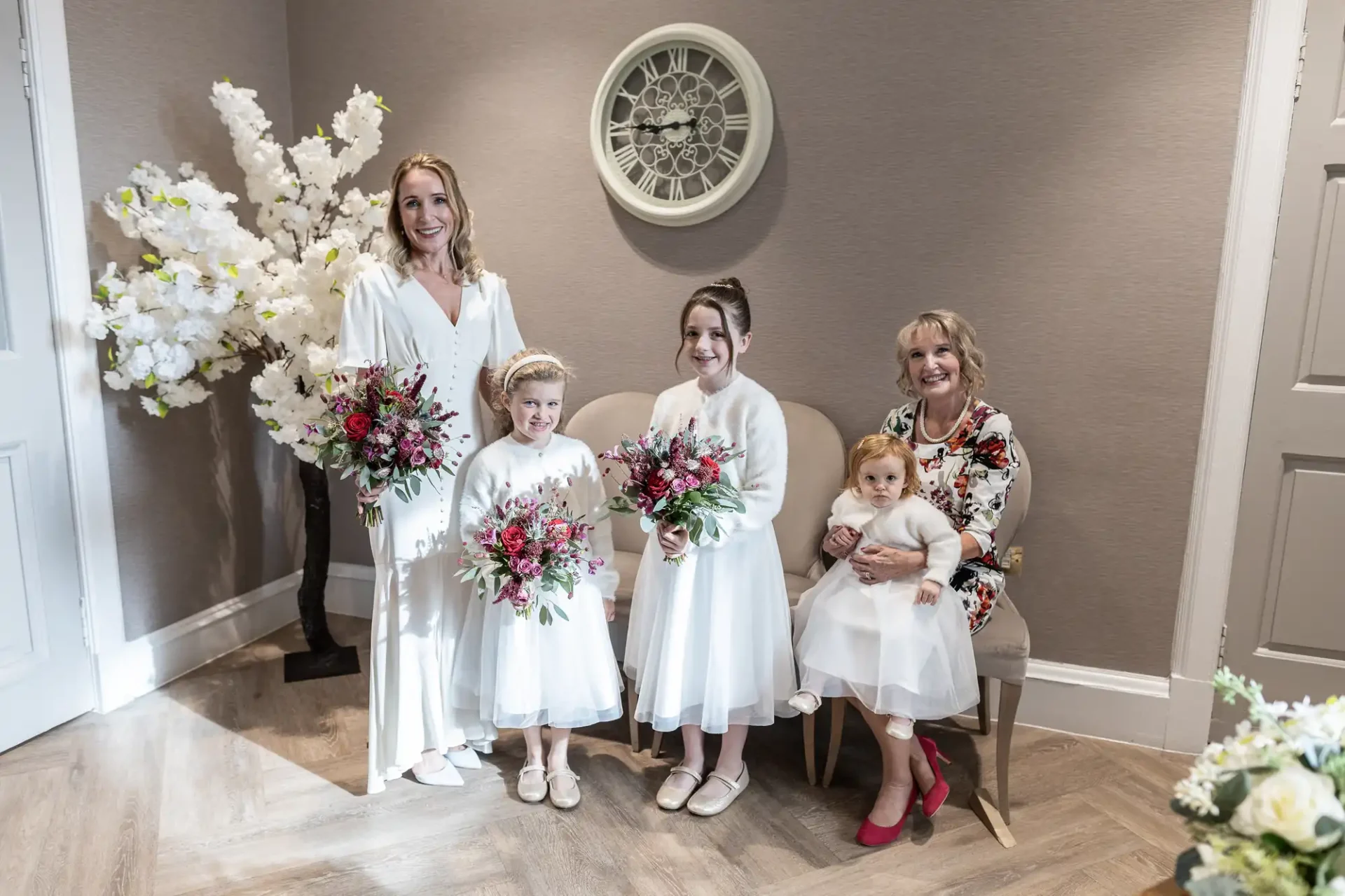 A group of five people in formal attire pose indoors; three girls in white dresses, an adult in a white dress, and an adult in a floral dress. Flowers and a clock adorn the background.