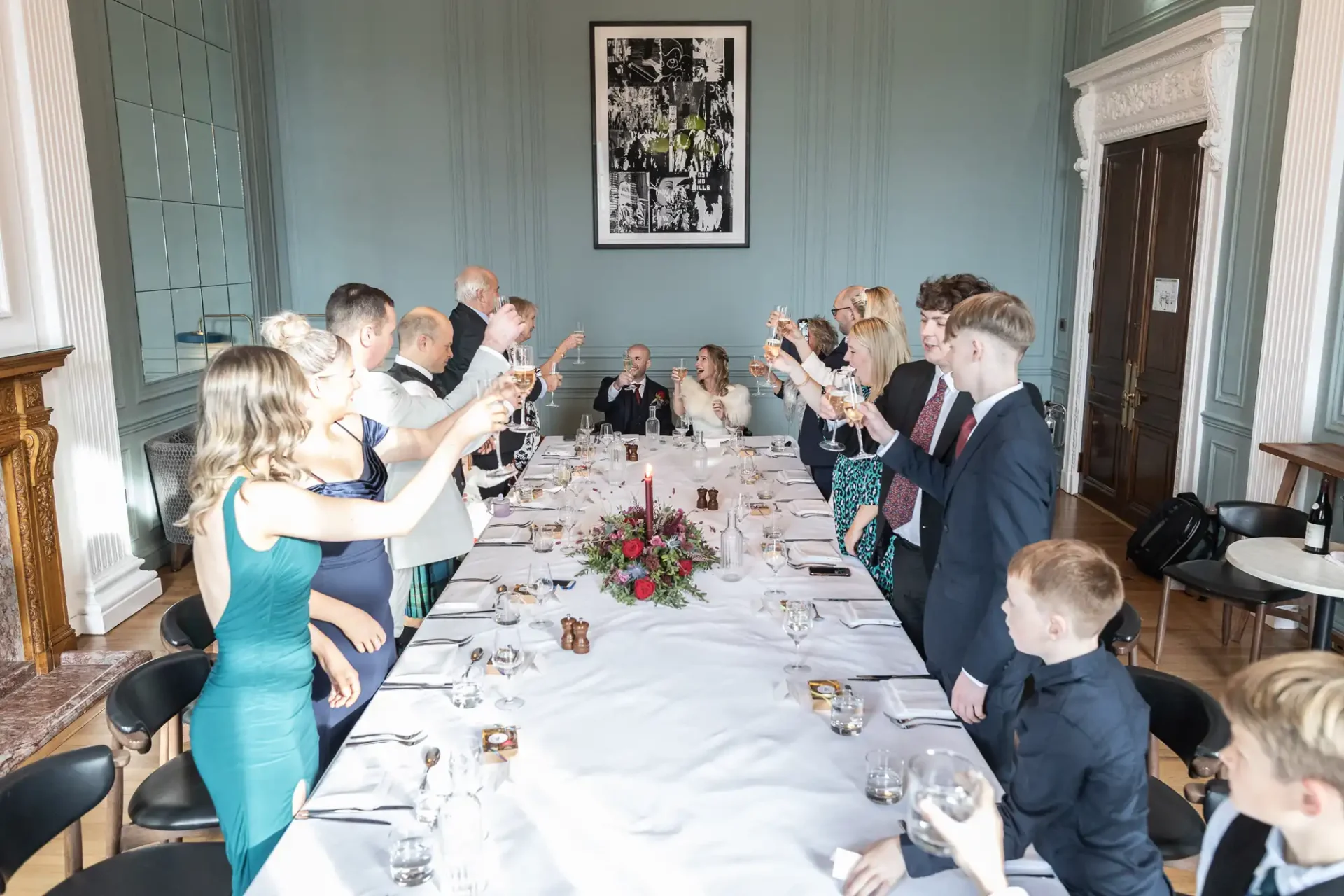 Group of people standing and raising glasses around a long dining table, set with white tablecloth and floral centerpiece, in an elegant room.