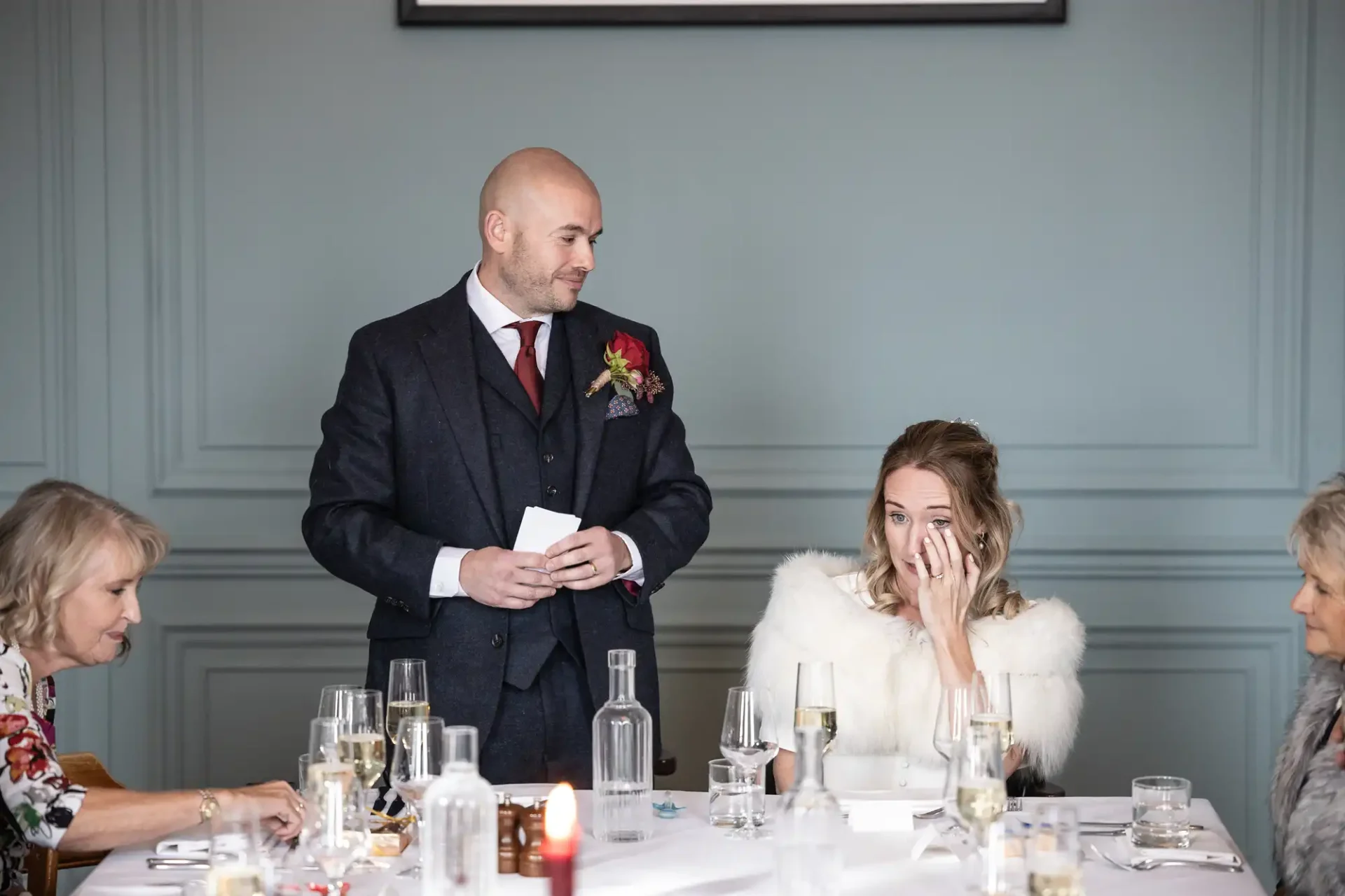 A man in a suit gives a speech at a wedding reception. A woman in a white fur shawl sits beside him, appearing emotional. Two older women sit at the table, one looking on.