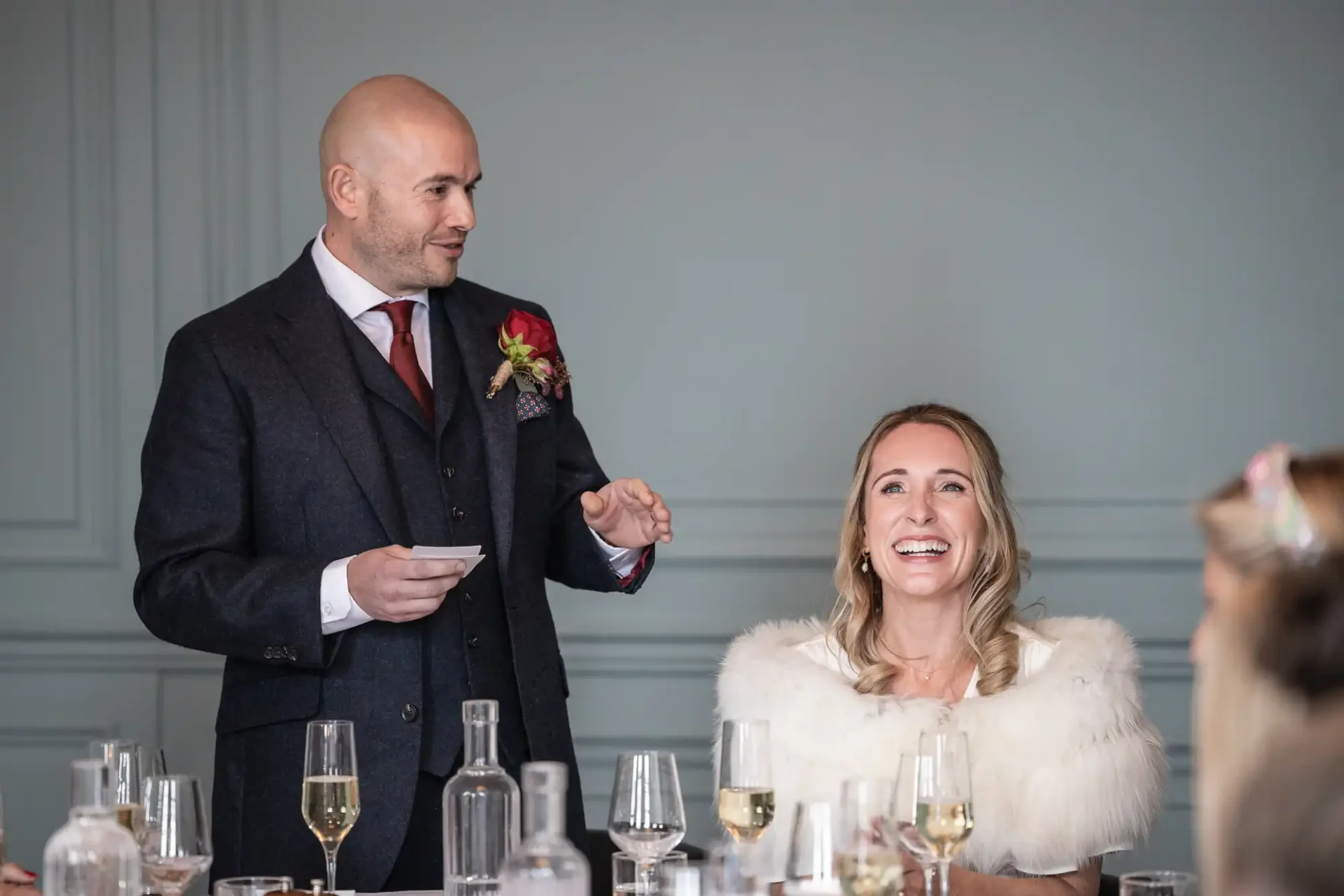 A man in a suit gives a speech next to a smiling woman in a white wrap seated at a table with glasses and bottles.