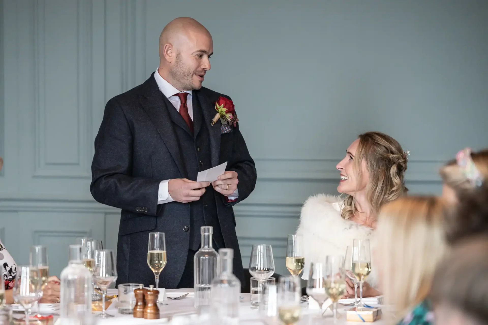 A man in a suit gives a speech at a formal dinner while a woman in a light-colored outfit smiles at him. They are surrounded by glasses and bottles on a white tablecloth.