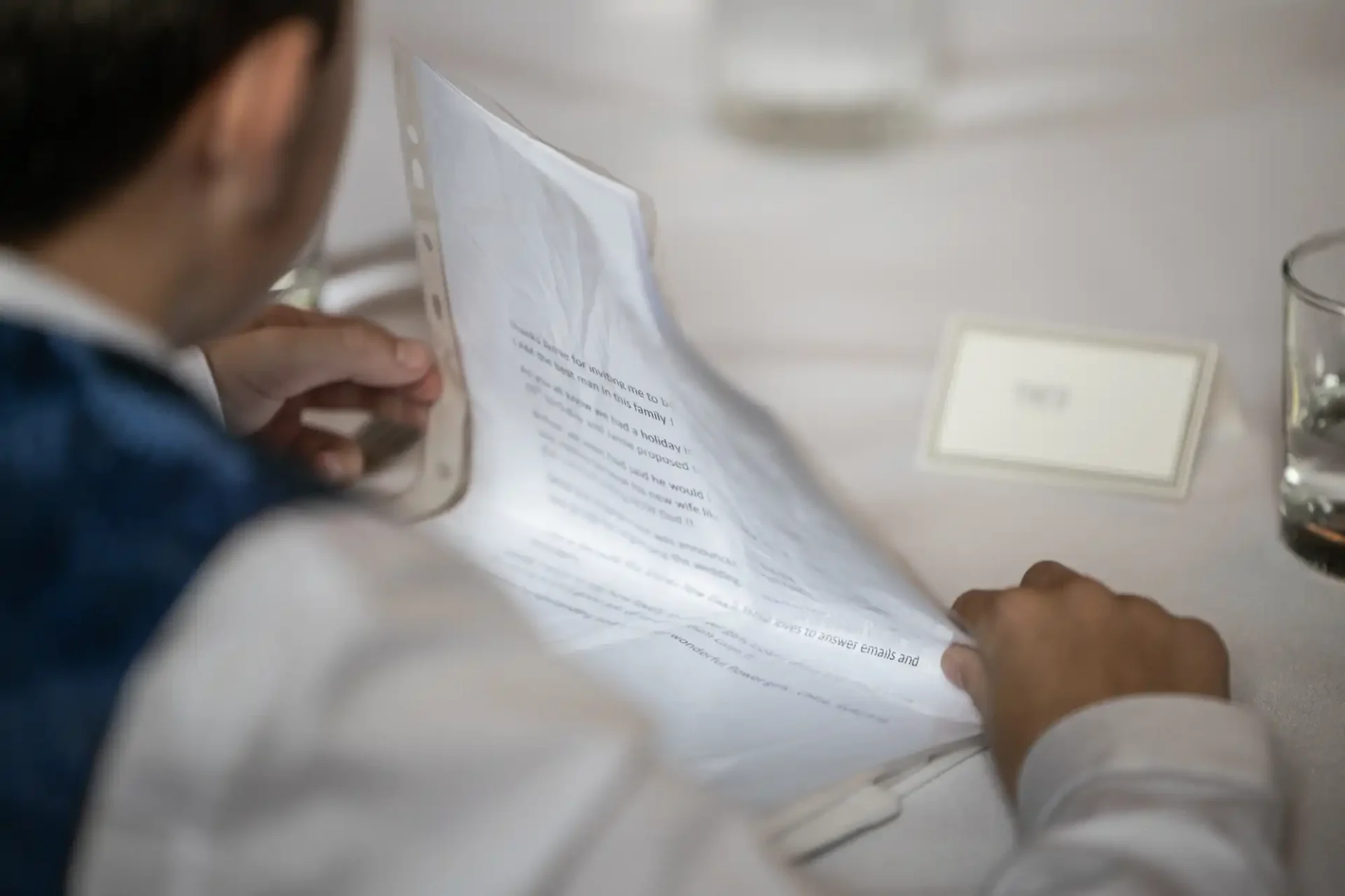 A person in a white shirt is sitting at a table, reading a slightly crumpled sheet of paper. Nearby are a glass of water and a small, blank name card.