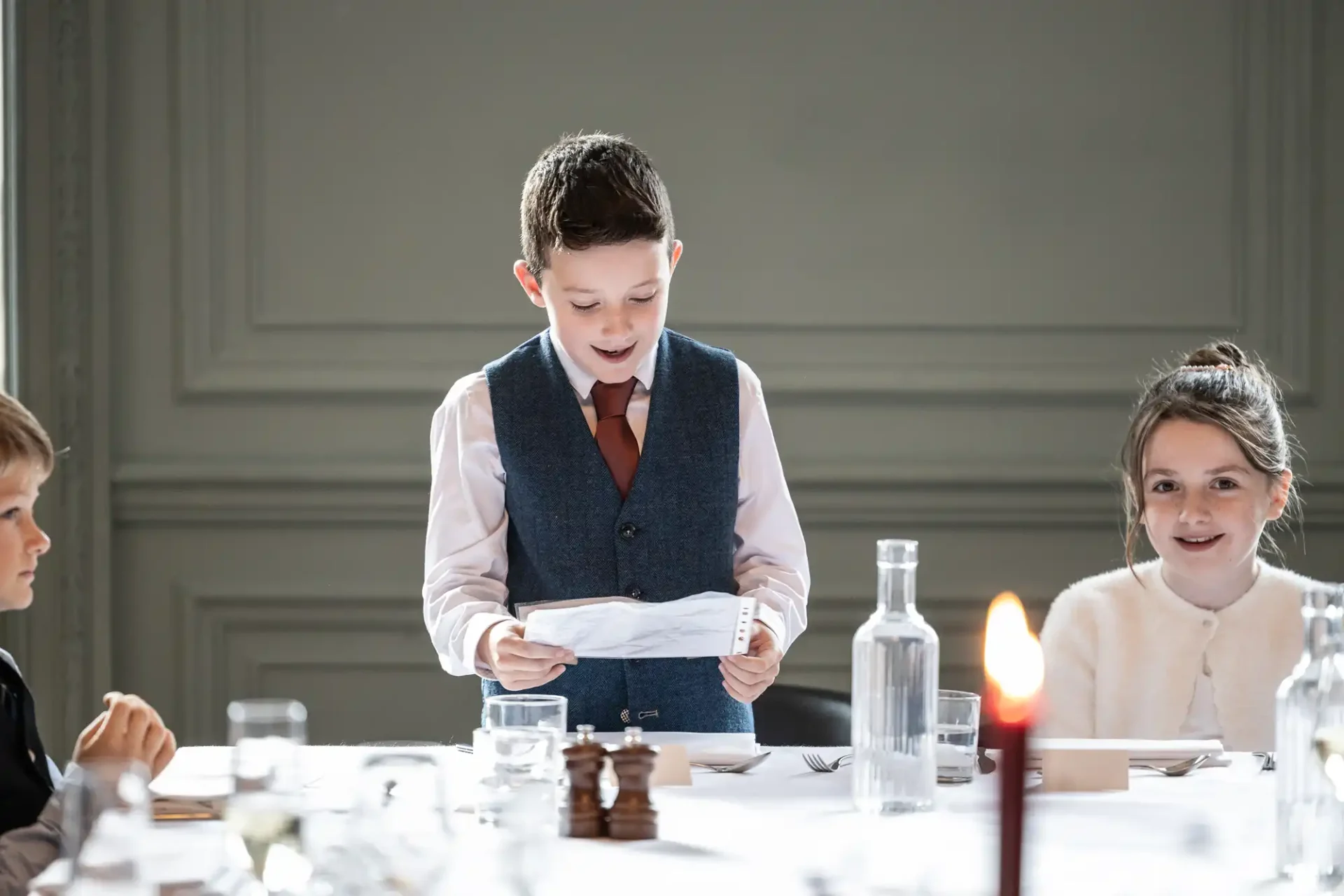 A young boy in a vest reads from a paper at a table set with glassware and a candlestick, while other children listen.