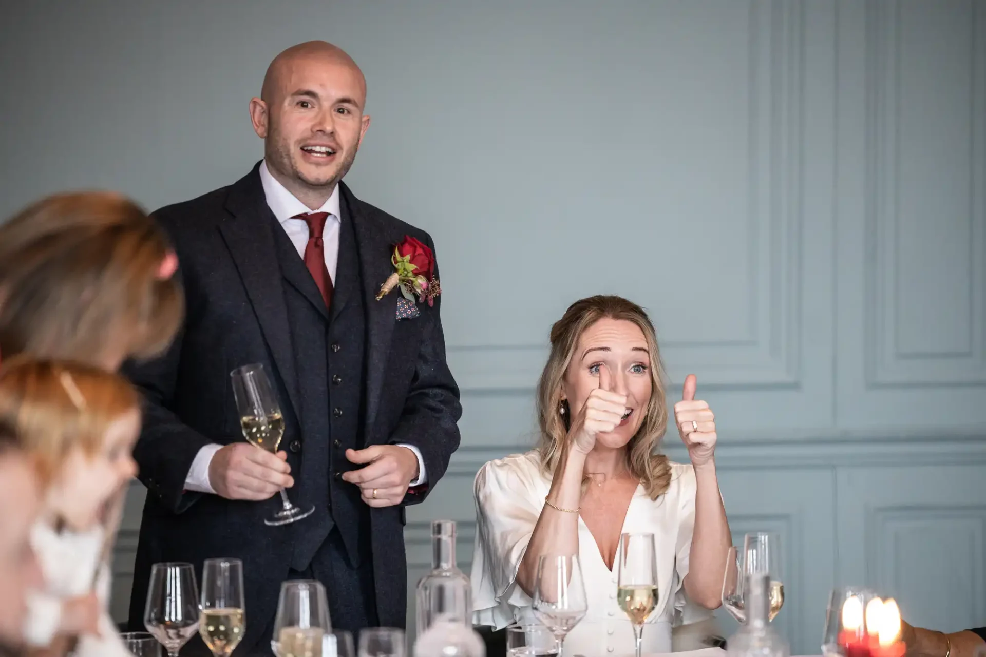 A man in a suit gives a speech holding a wine glass, while a woman beside him shows two thumbs up. Two children are partially visible in the foreground.