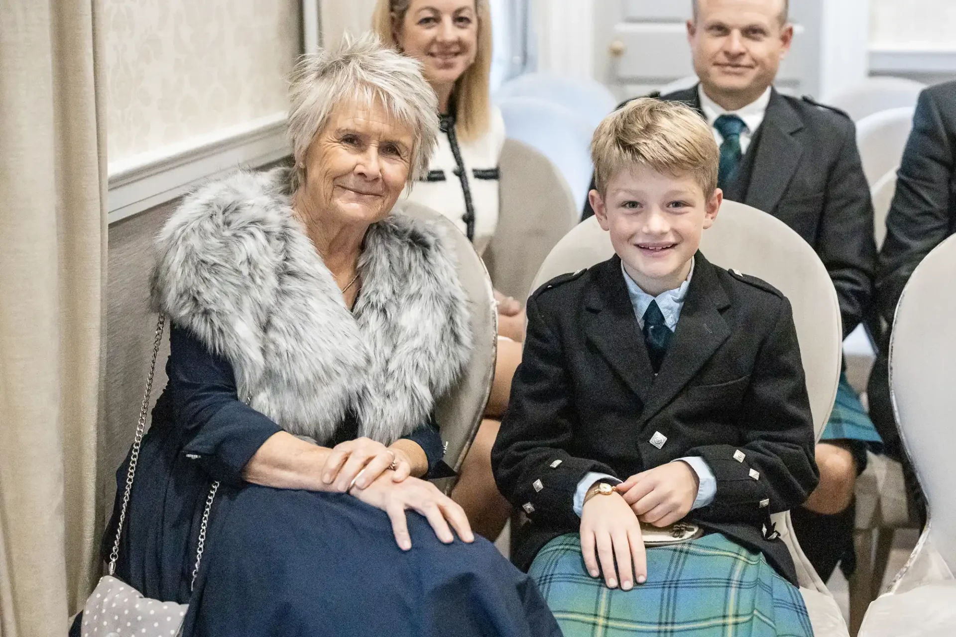 An elderly woman and a boy in Scottish attire sit together on chairs, surrounded by others in a room.