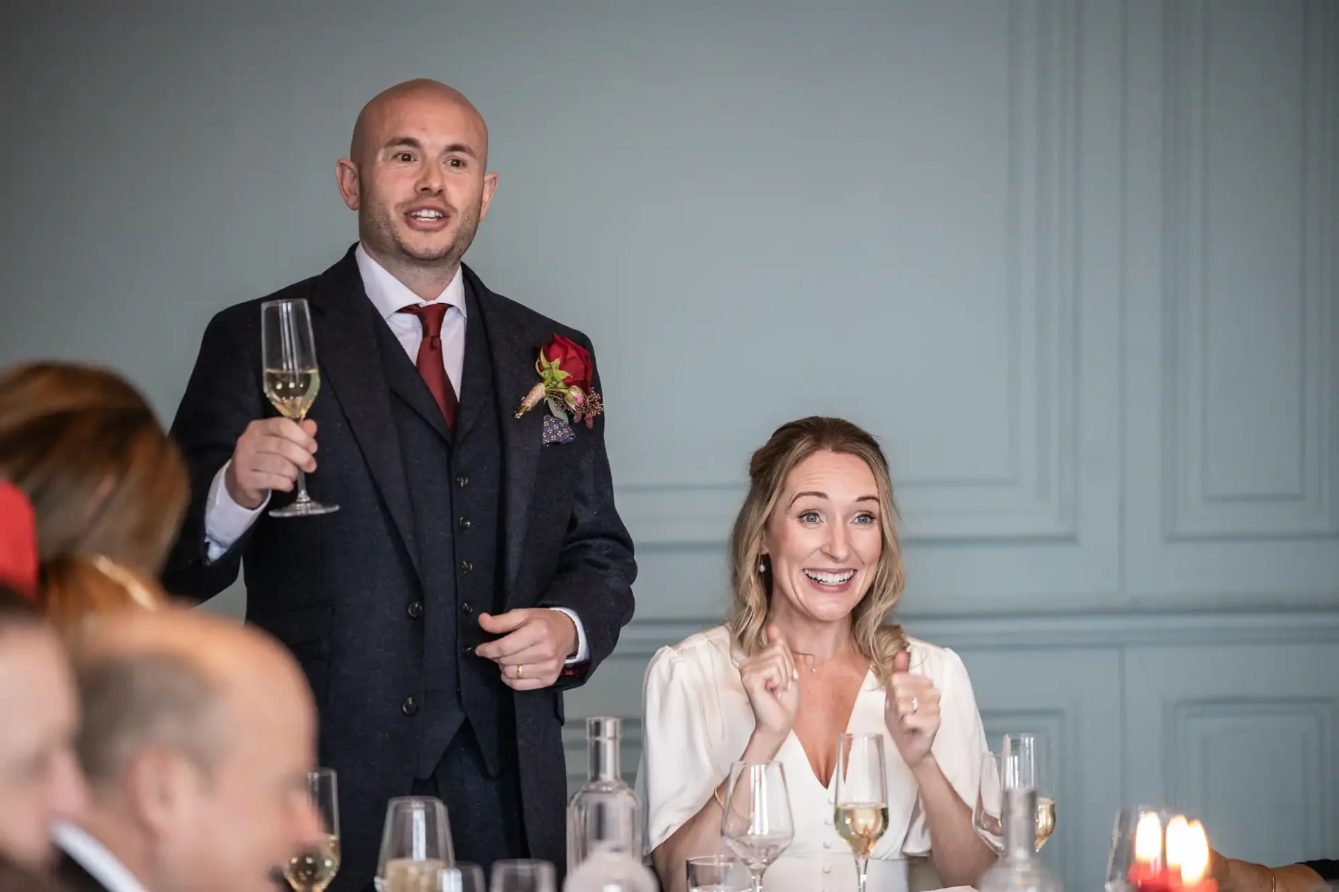 A man in a suit raises a glass while standing. A seated woman smiles joyfully with her hands slightly raised. Glasses and bottles are on the table in front of them.