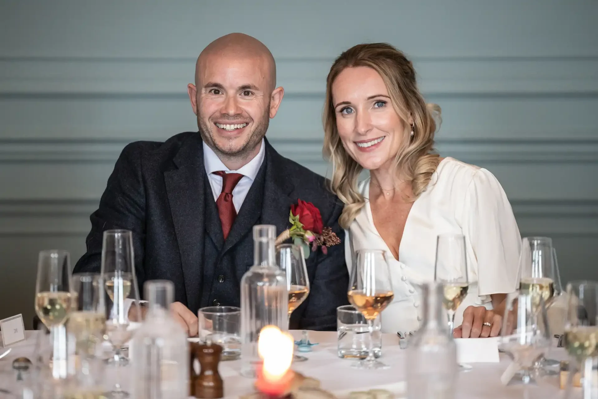 A man and woman sitting at a dining table with wine glasses, candles, and bottled water, smiling at the camera.