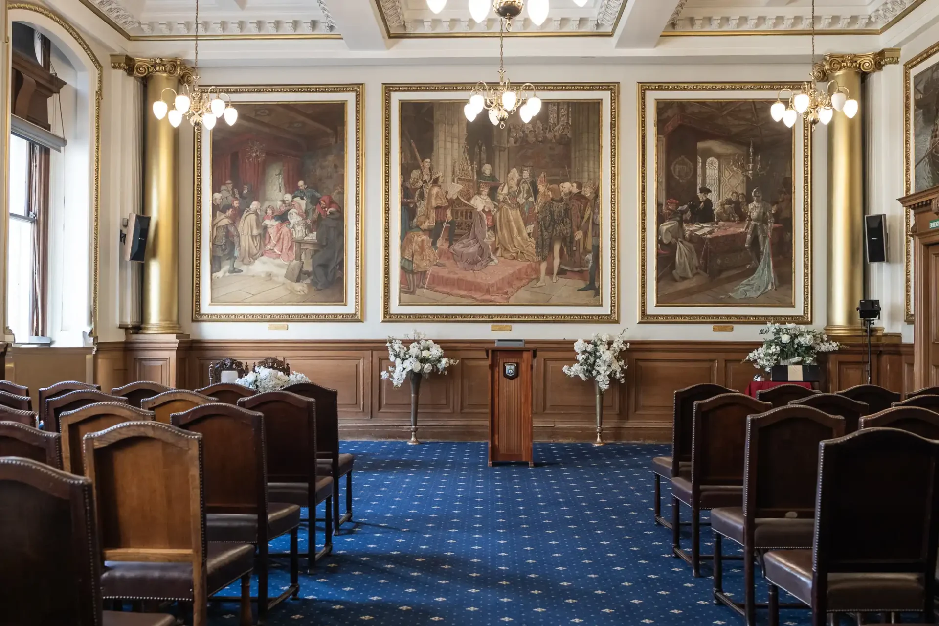 A room with wooden chairs, a podium, and three large paintings on the wall. Chandeliers hang from the ceiling, and floral arrangements are placed beside the podium.